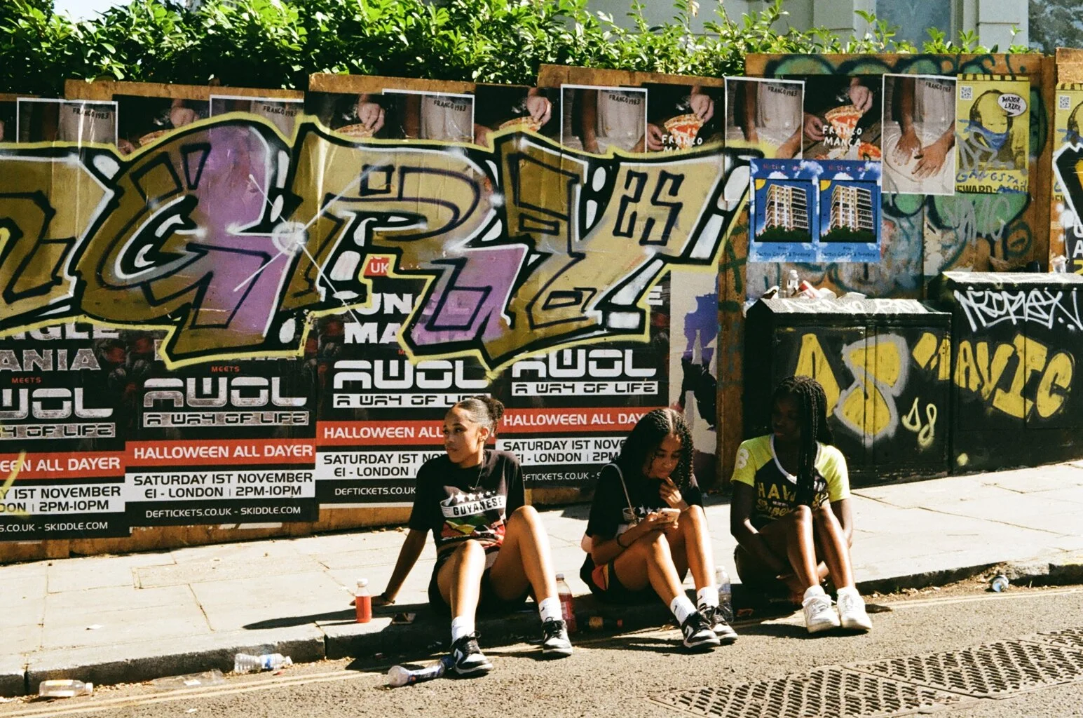 Three young women sit on the sidewalk against a graffiti-covered wall, with posters advertising an event called 'Augl'.