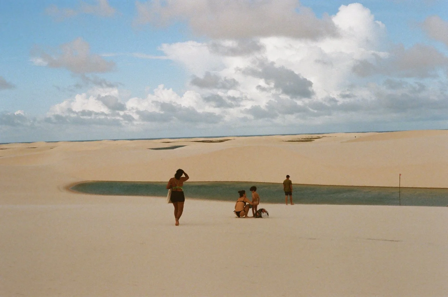 Beach with white sand dunes, a small lake, and four people relaxing and taking photos under cloudy sky.