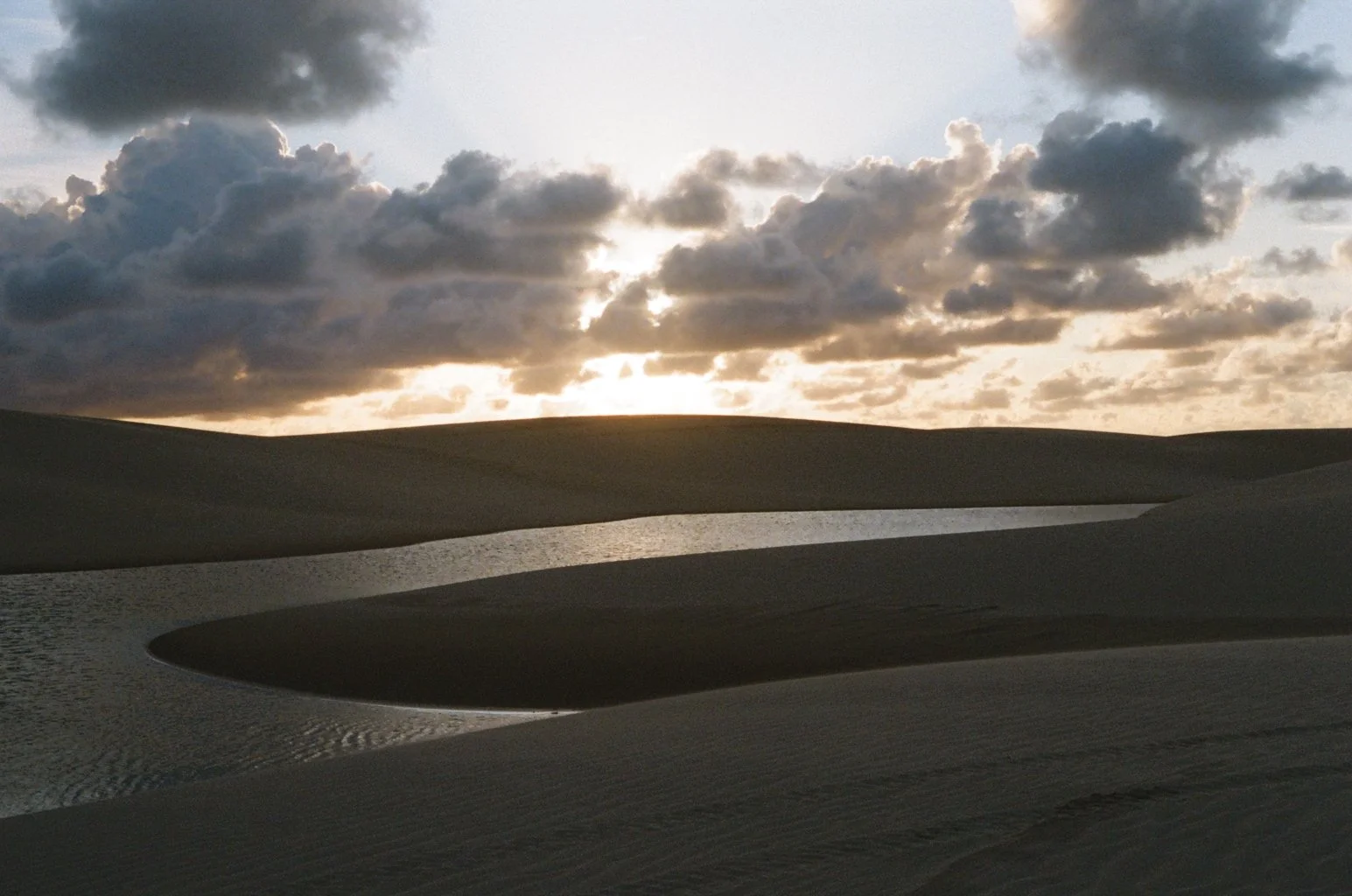 Sand dunes with a water body underneath a cloudy sky at sunset or sunrise.