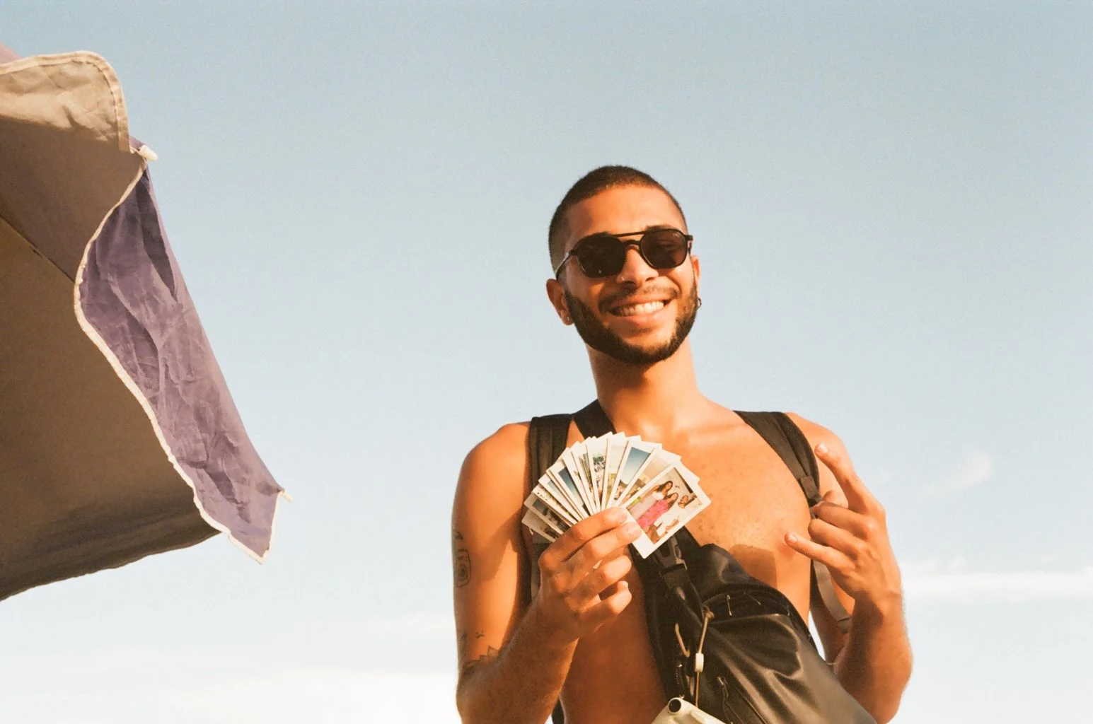 A smiling man wearing sunglasses, a backpack, and a crossbody bag, holding a fan of postcards or photographs, making the shaka gesture with his right hand, outdoors under a clear sky.
