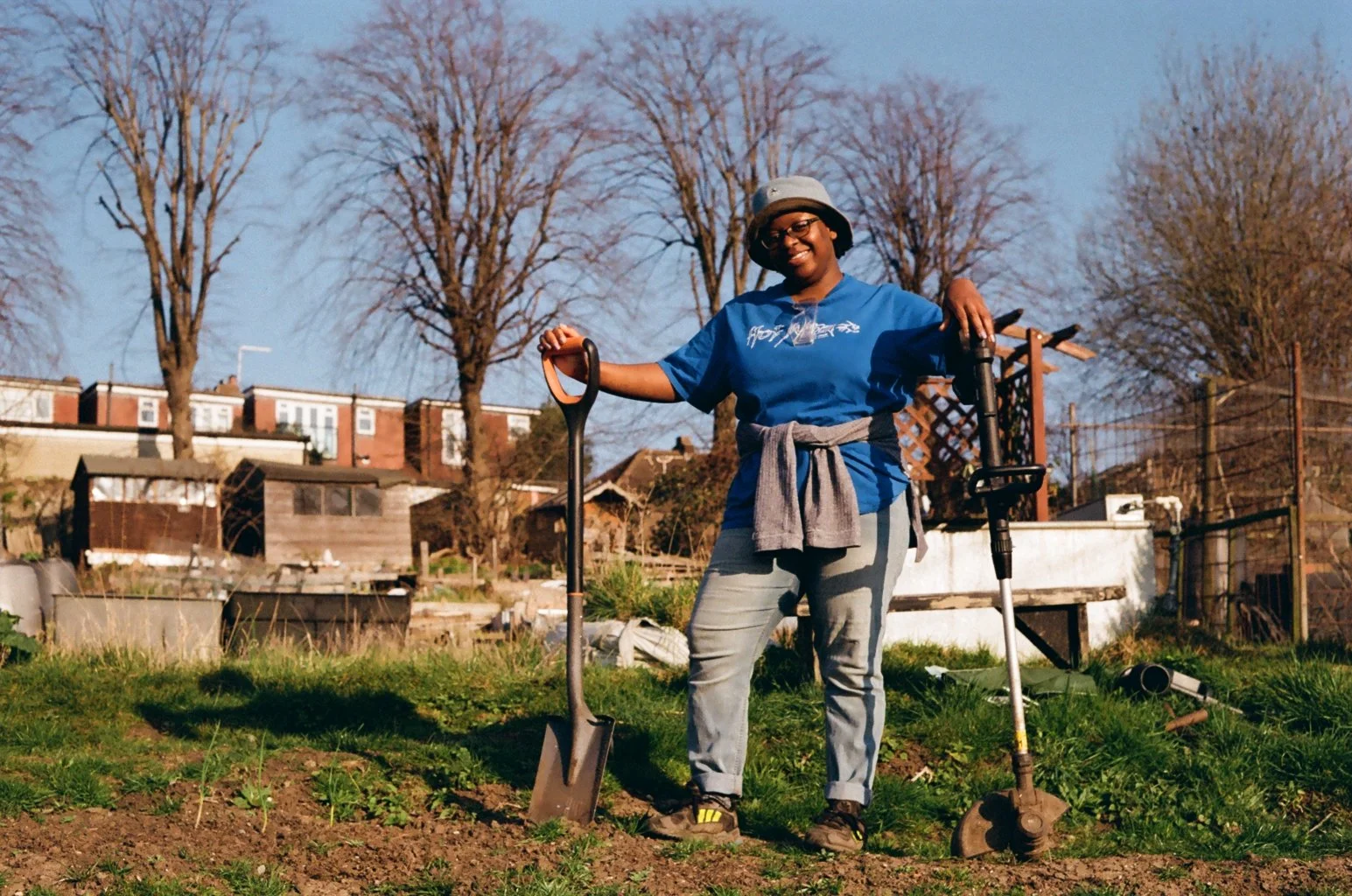 A woman standing outdoors in a garden, holding a shovel and a power tool, smiling. She is wearing a blue t-shirt, gray pants, and a hat, with a gray sweater tied around her waist. In the background, there are trees, residential buildings, and garden 