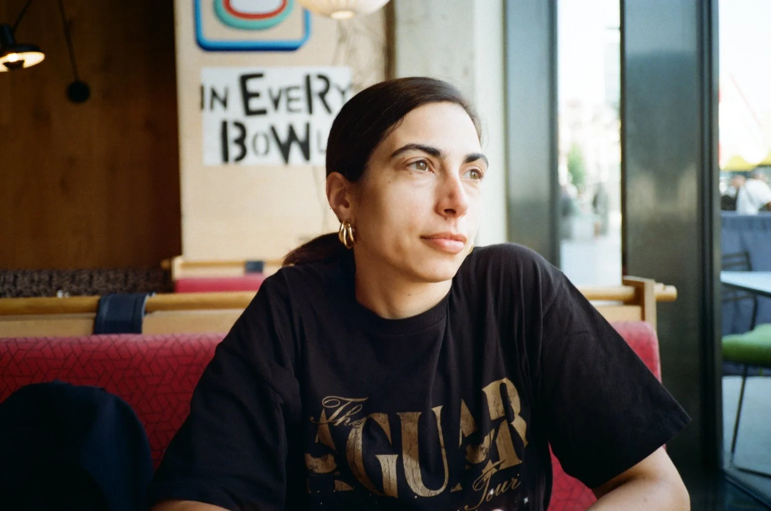 A woman with dark hair and gold hoop earrings sitting in a restaurant, looking out the window, with a sign that says 'IN EVERY BOWL' behind her.