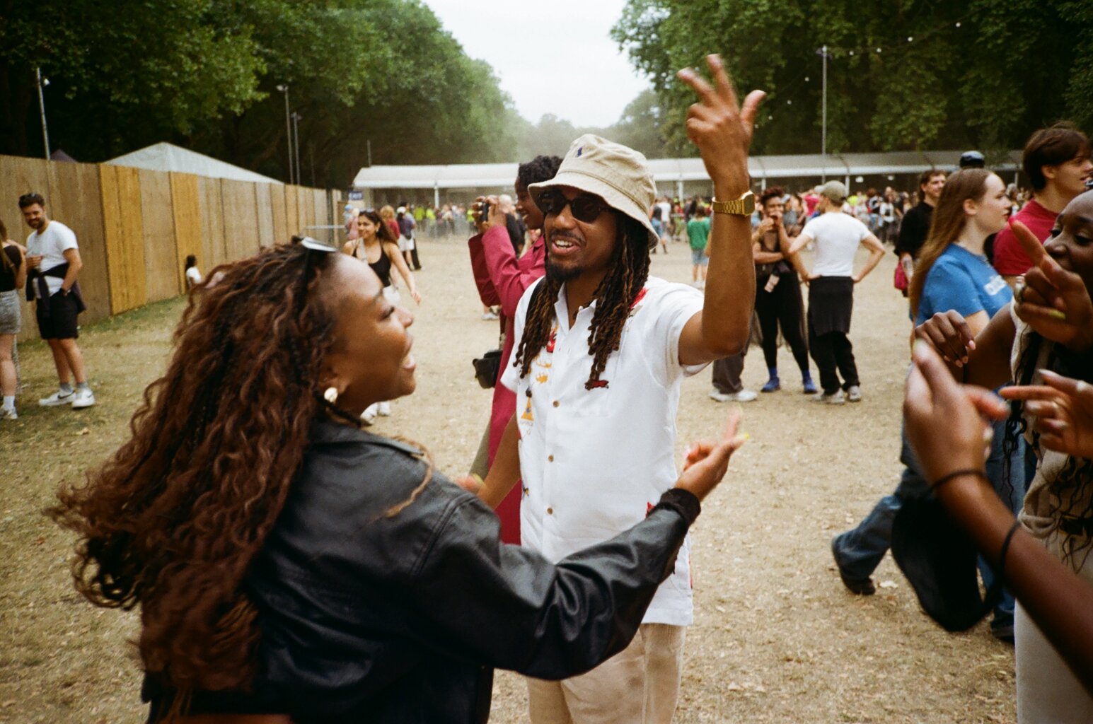 A Black man with dreadlocks wearing a hat and sunglasses is talking and gesturing to a woman with curly hair and a leather jacket at an outdoor event, with other people in the background.