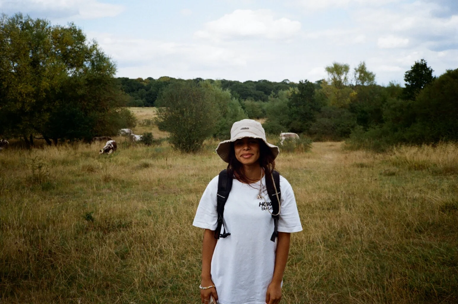 A woman standing in a grassy field with trees and cows in the background under a partly cloudy sky.