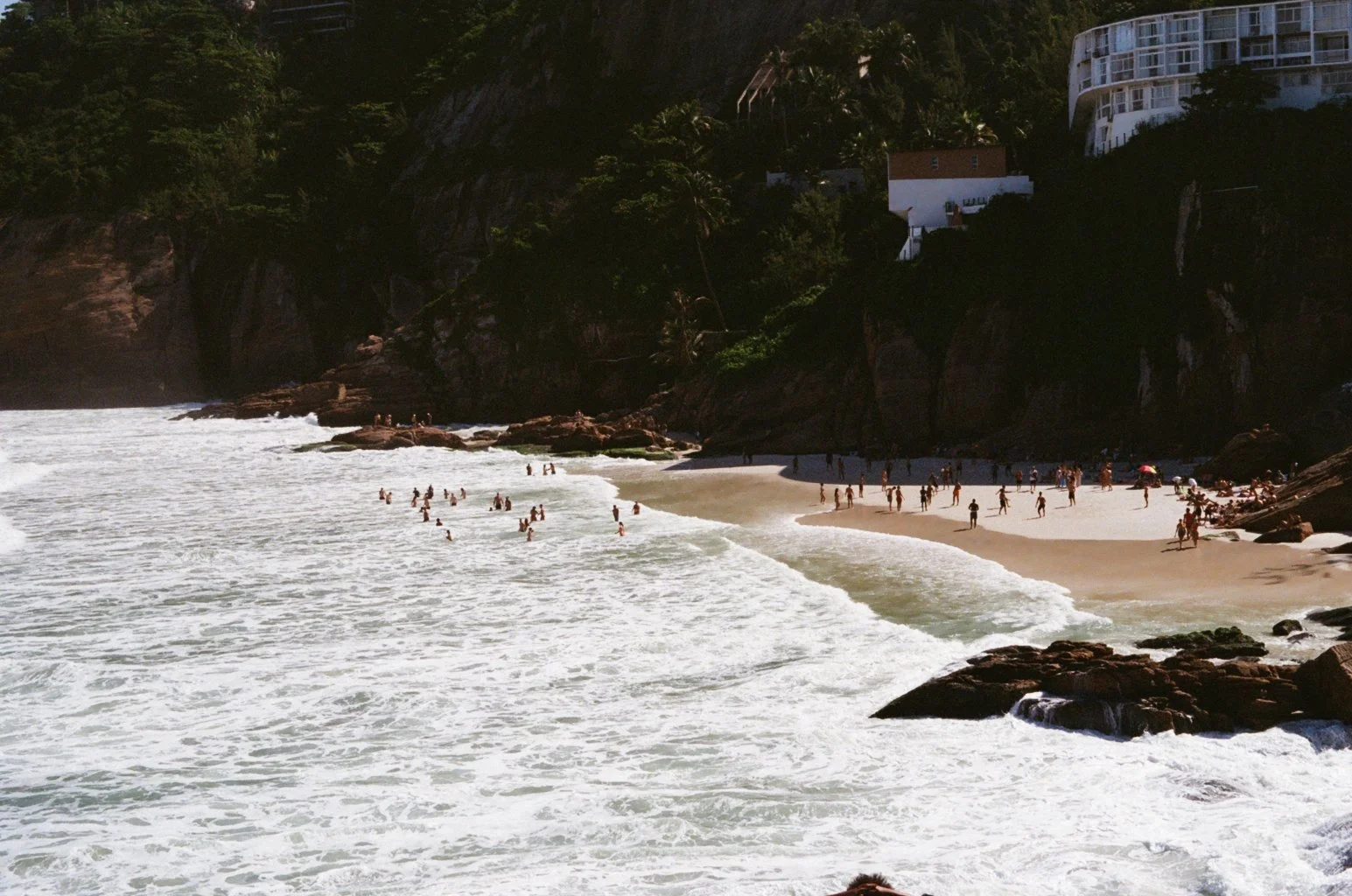 Beach scene with people swimming and walking along the sandy shore, rocky coastline, and cliffside with buildings in the background.