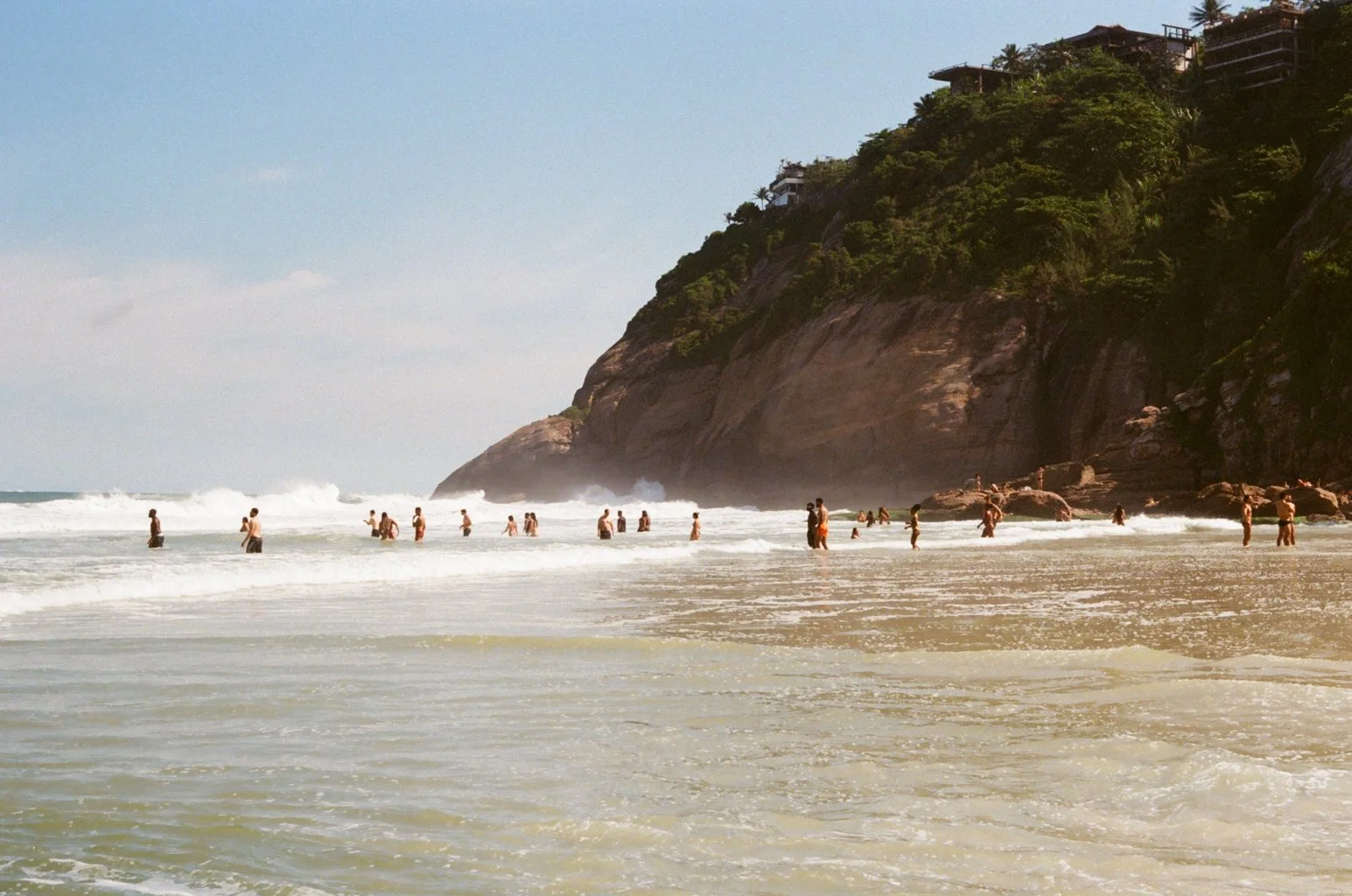 People enjoying the beach with a large rocky cliff and green vegetation in the background, clear sky, and waves crashing onto the shore.