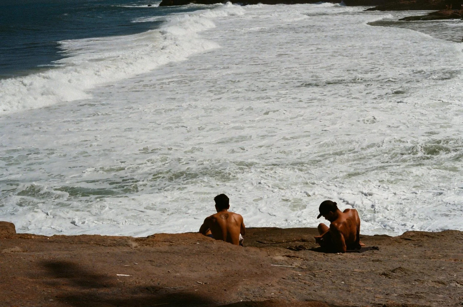 Two men sitting on a rocky beach facing the ocean waves, one wearing a cap and the other with short hair, both shirtless.