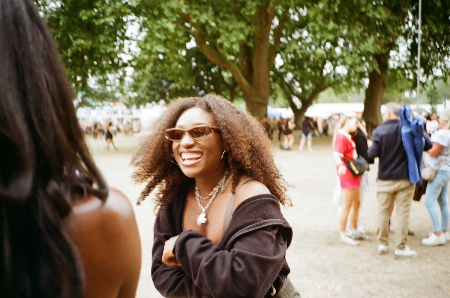 A Black woman with curly hair wearing sunglasses, a black jacket, and layered necklaces, smiling and talking to someone at an outdoor event with trees and other people in the background.