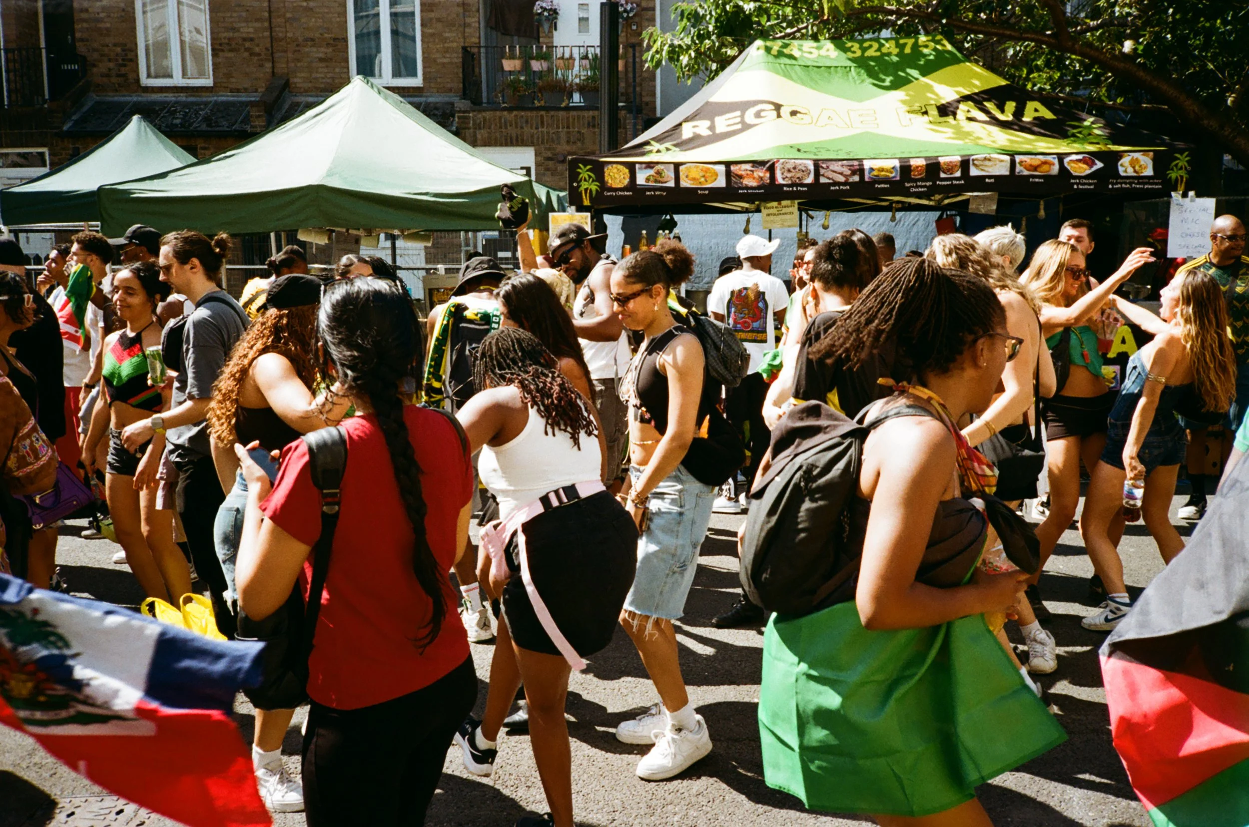 Crowd of diverse people celebrating an outdoor event with a green and yellow canopy and food stand in the background, some dancing and others socializing.