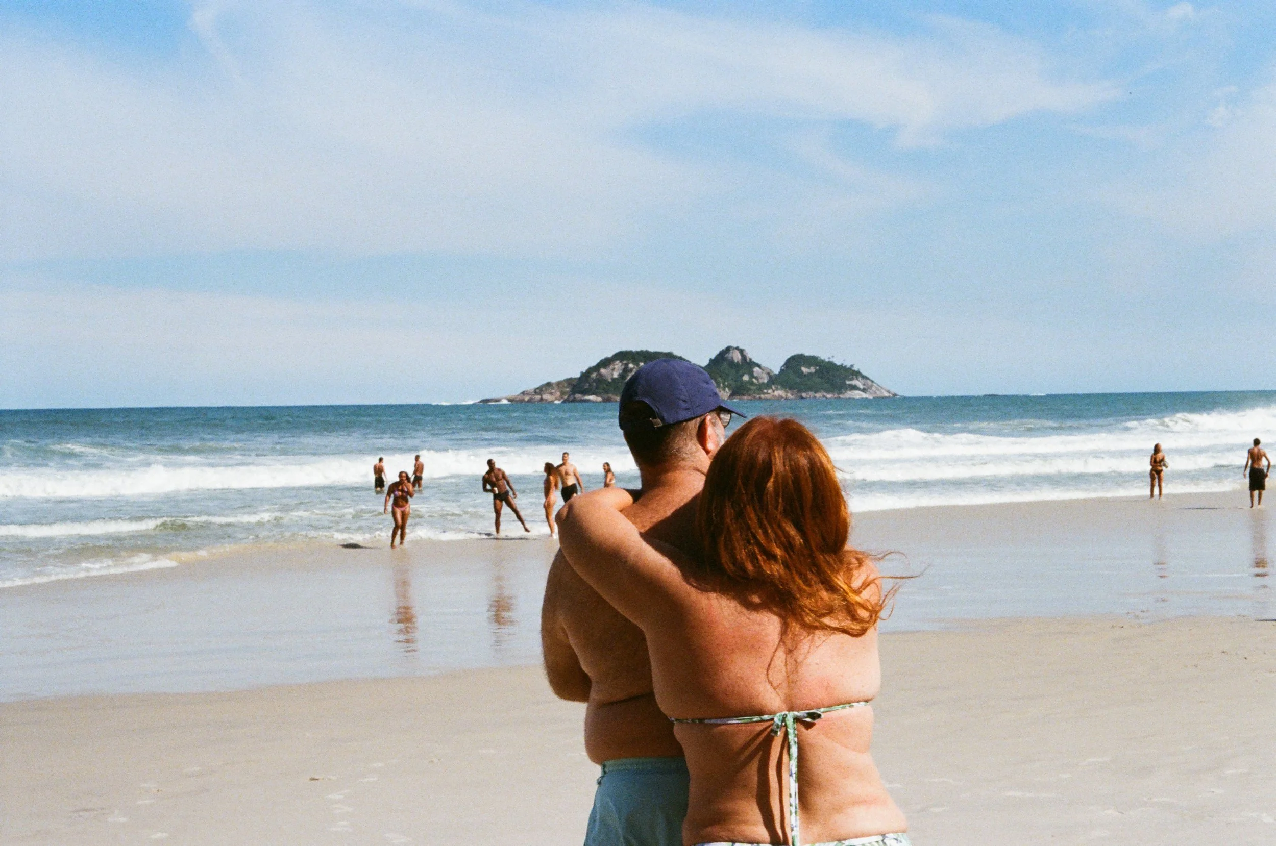 Couple embracing on a beach with people wading in the ocean, and rocky islands in the background under a partly cloudy sky.