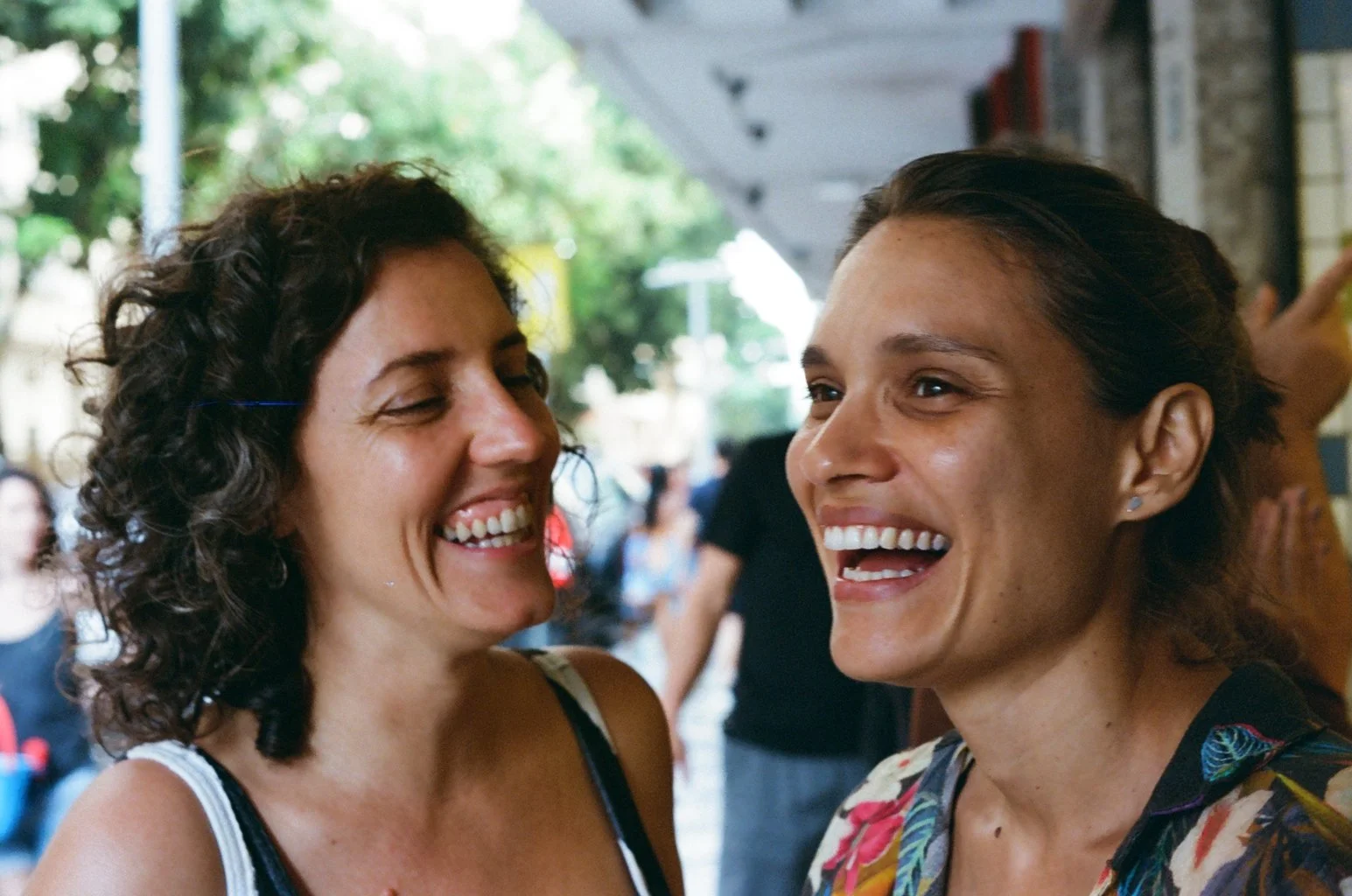 Two women smiling and laughing together outdoors, with trees and people in the background.