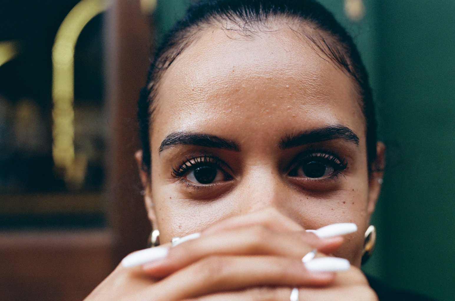 A close-up of a young woman with dark skin, dark hair, and brown eyes covering her mouth with her hand, showing white nail polish, in an indoor setting with a blurred background.