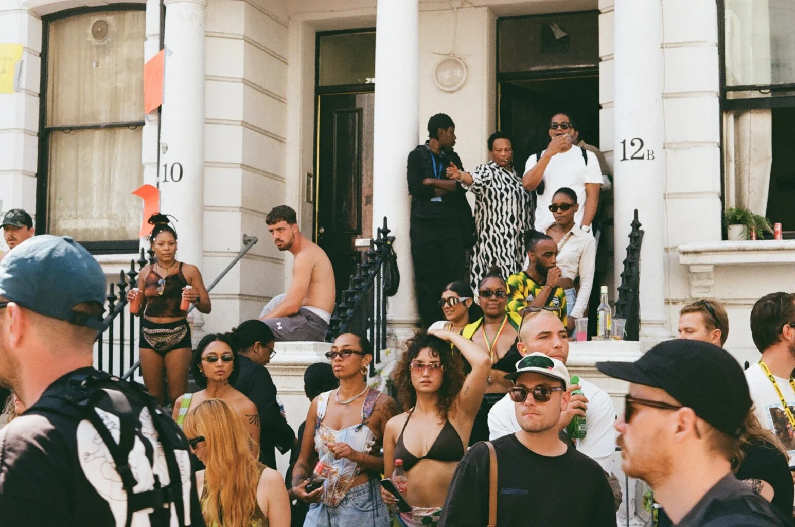 Crowd of diverse young adults gathering outdoors on the steps of a building, some wearing sunglasses and casual summer clothes, engaging in conversation and holding drinks.