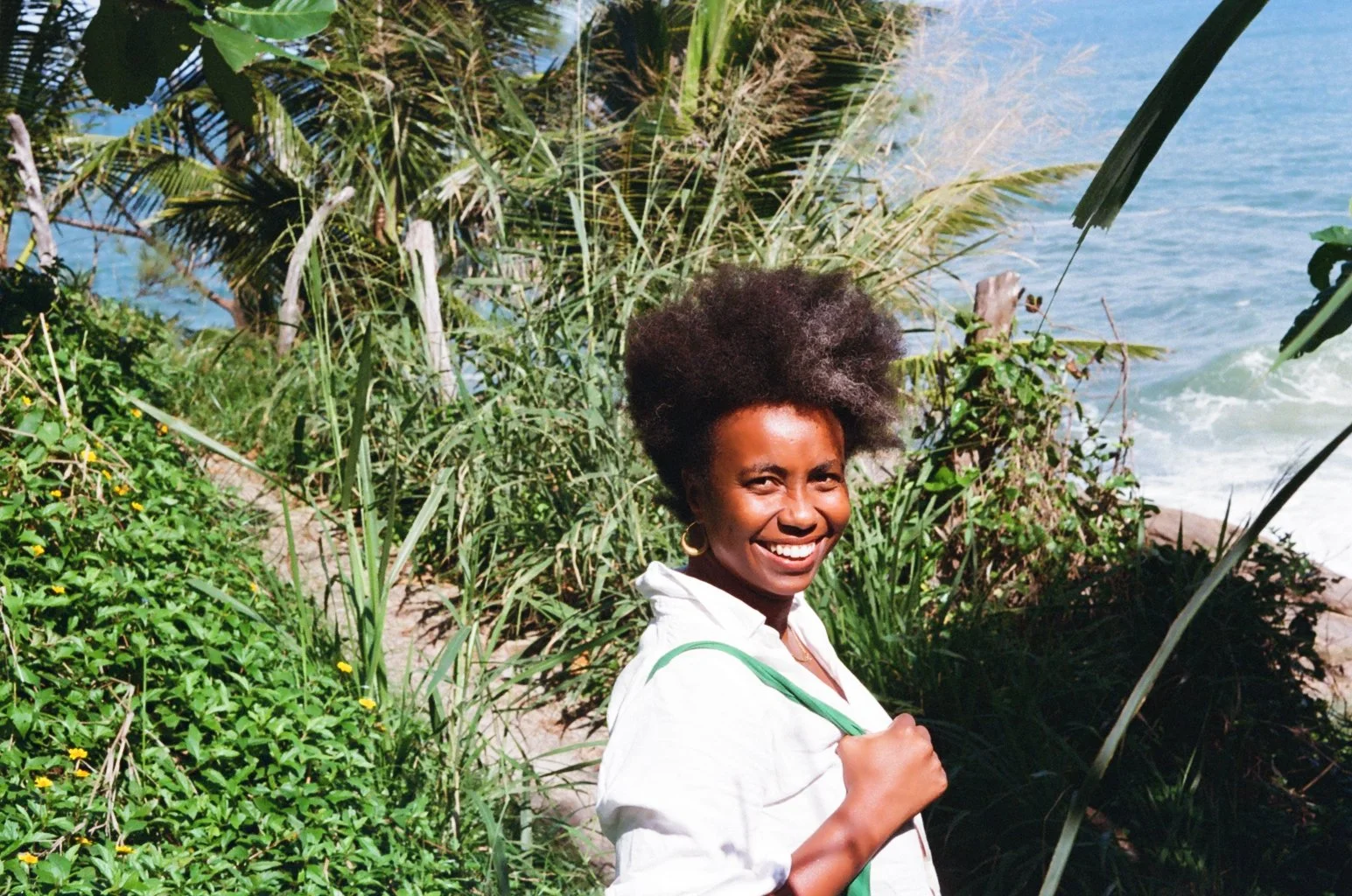 A smiling woman with glowing brown skin and an afro hairstyle, wearing a white shirt and gold hoop earrings, standing on a beach with lush green tropical plants and a blue ocean in the background.