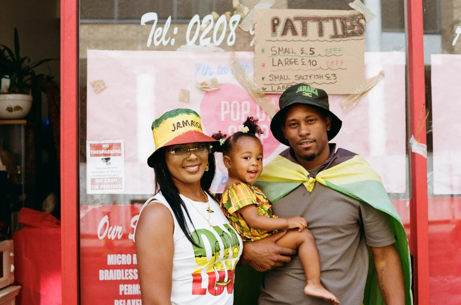 A family of three celebrating Jamaican culture, with the mother and father wearing Jamaican hats, and the child draped in a Jamaican flag, standing in front of a storefront with Jamaican-themed signs and posters.