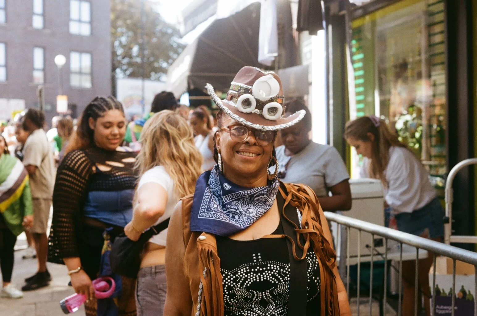 A woman wearing a decorated cowboy hat with buttons, a brown fringed jacket, glasses, and a blue bandana, smiling at an outdoor event with a crowd of diverse people in the background.