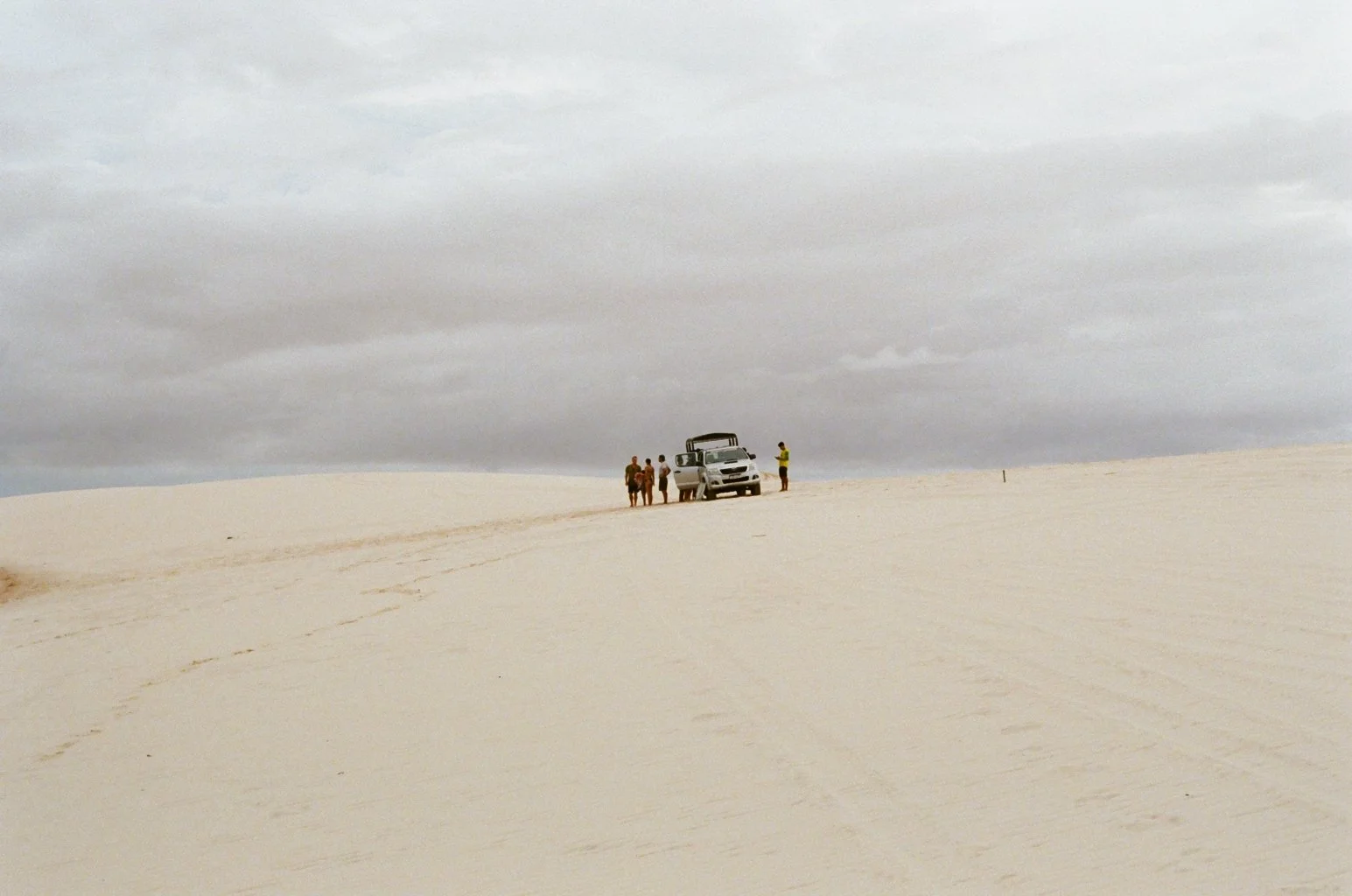 A group of people and a vehicle on a sandy dune under a cloudy sky.