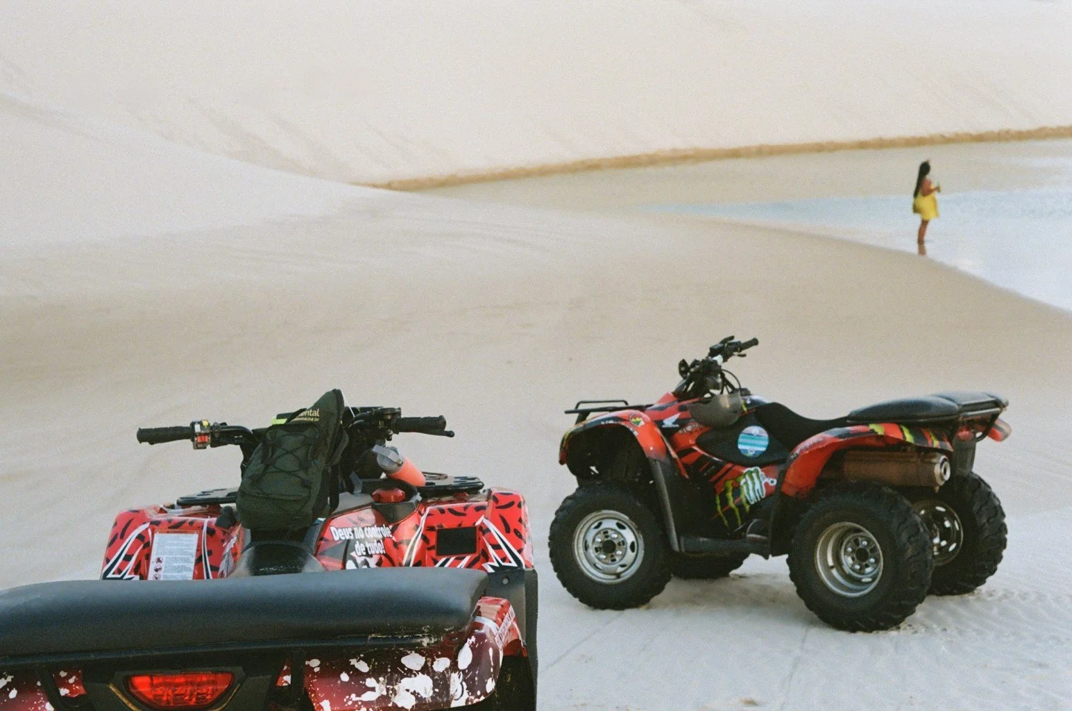 Two all-terrain vehicles parked on white sandy desert with a woman in yellow dress standing nearby.