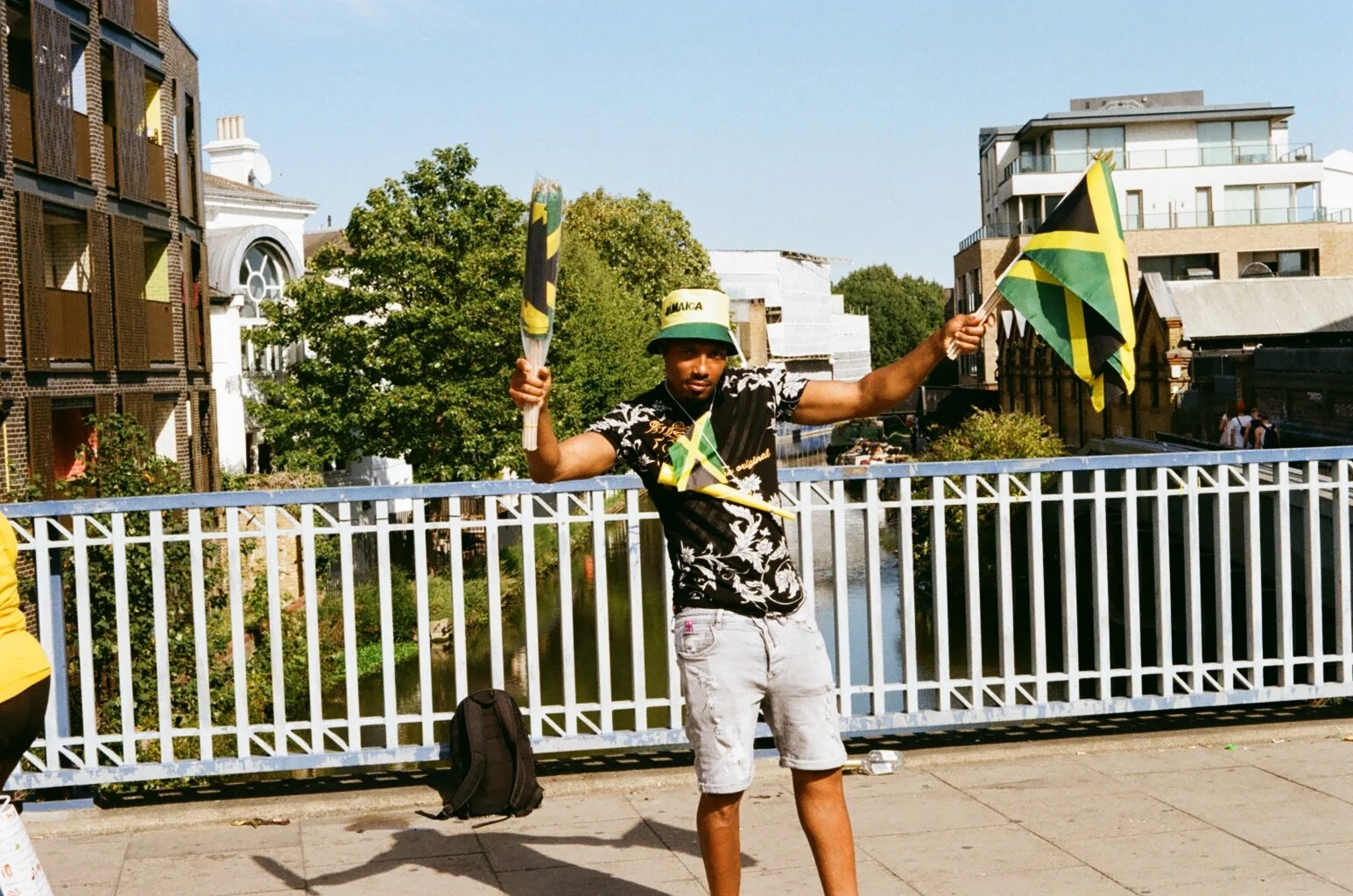 Young man standing near a bridge holding Jamaican flags, wearing a hat that says "Jamaica", black shirt with floral design, and gray shorts, with buildings and trees in the background.