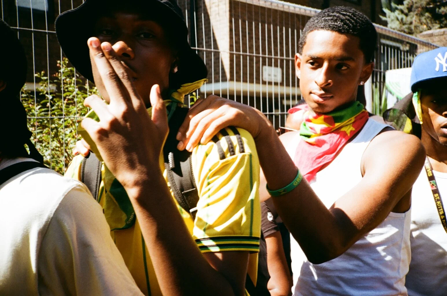 Three young men outdoors, one covering his mouth with his hand, wearing sports jerseys and bandanas, with a fence and greenery in the background.