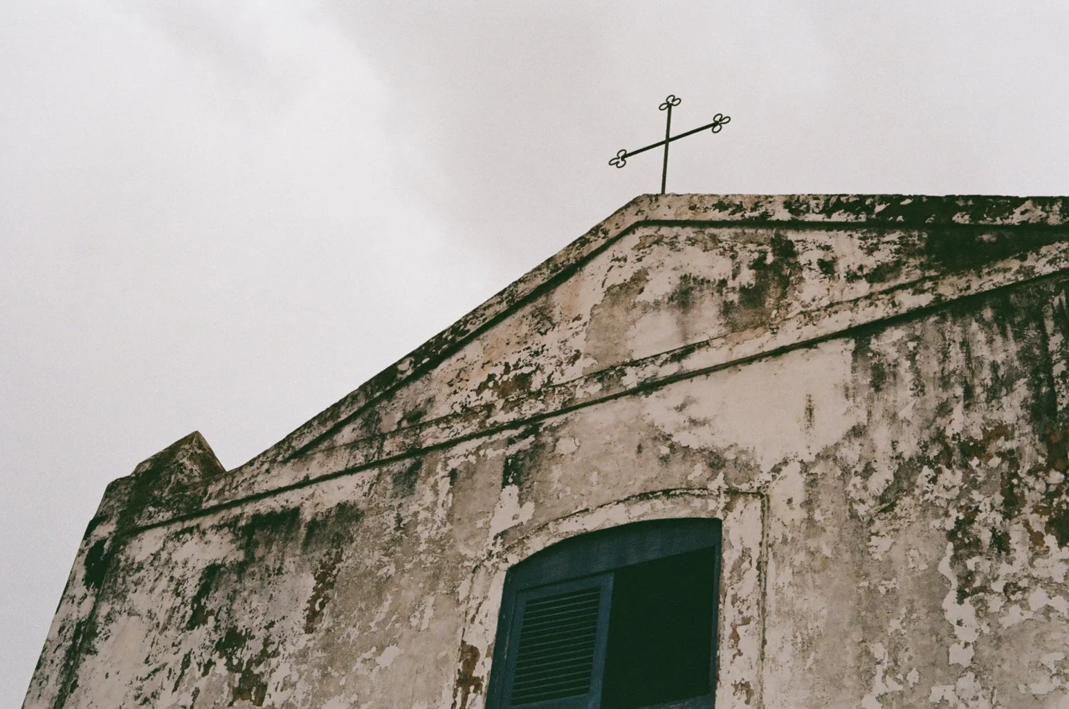 Close-up of an old, weathered church building with a cross on top and dark green window shutters.