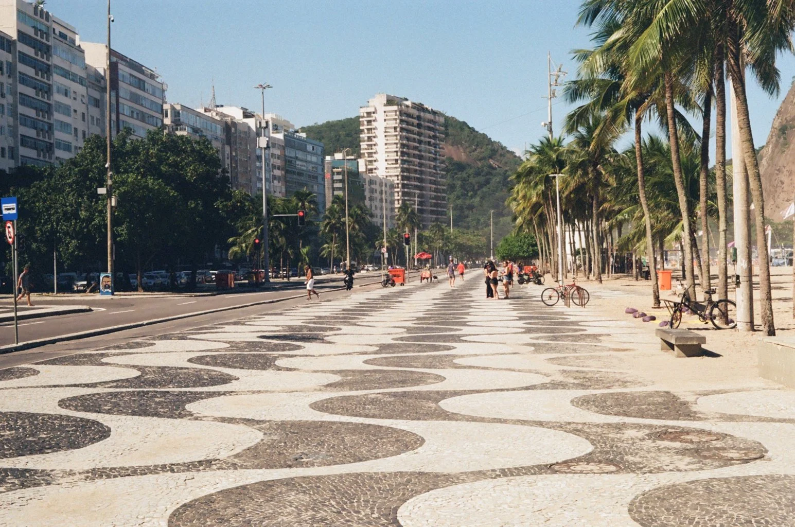 Beach promenade with palm trees, people walking, bicycles parked, high-rise buildings nearby, and a mountain in the background.