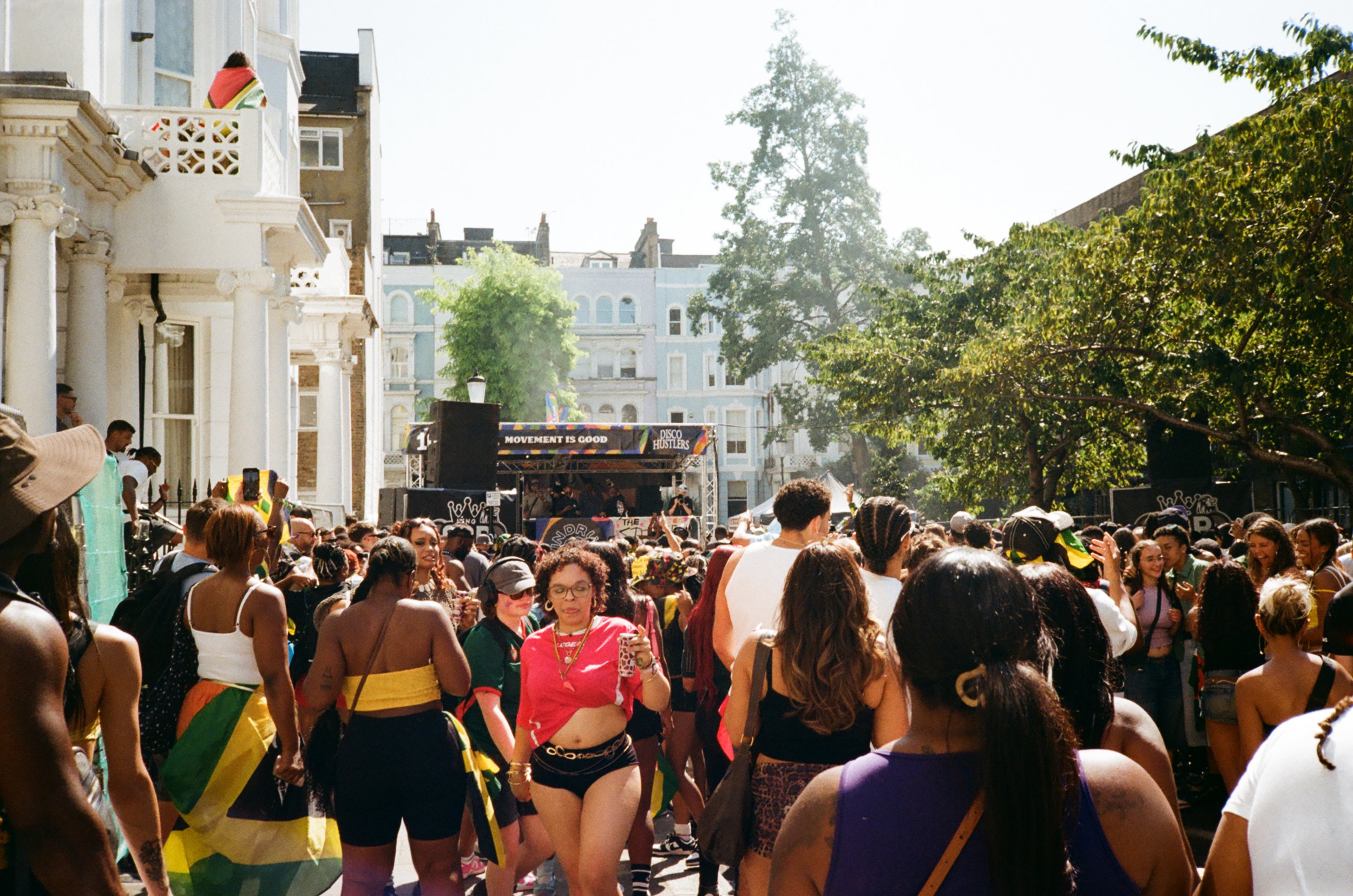 Crowd of people celebrating outdoors during daylight, with a stage in the background, trees, and residential buildings.