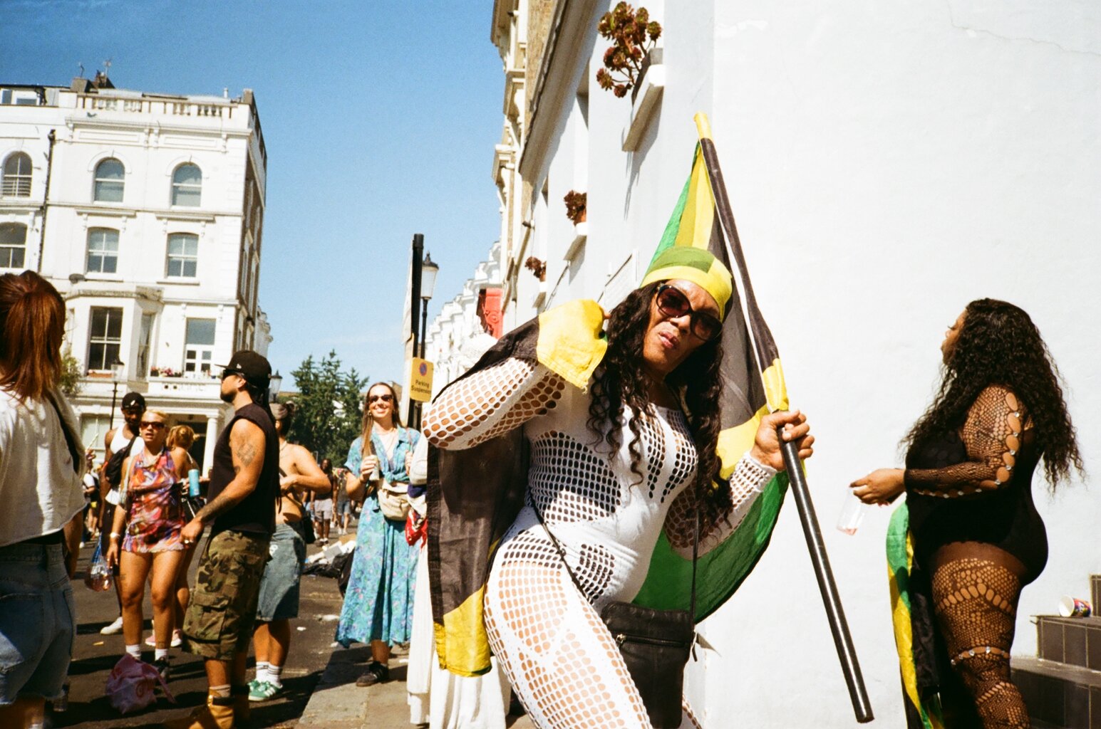 Woman with long curly hair wearing sunglasses, a yellow and green headscarf, and a black, yellow, and green flag while standing on a street with other people and white buildings in the background.