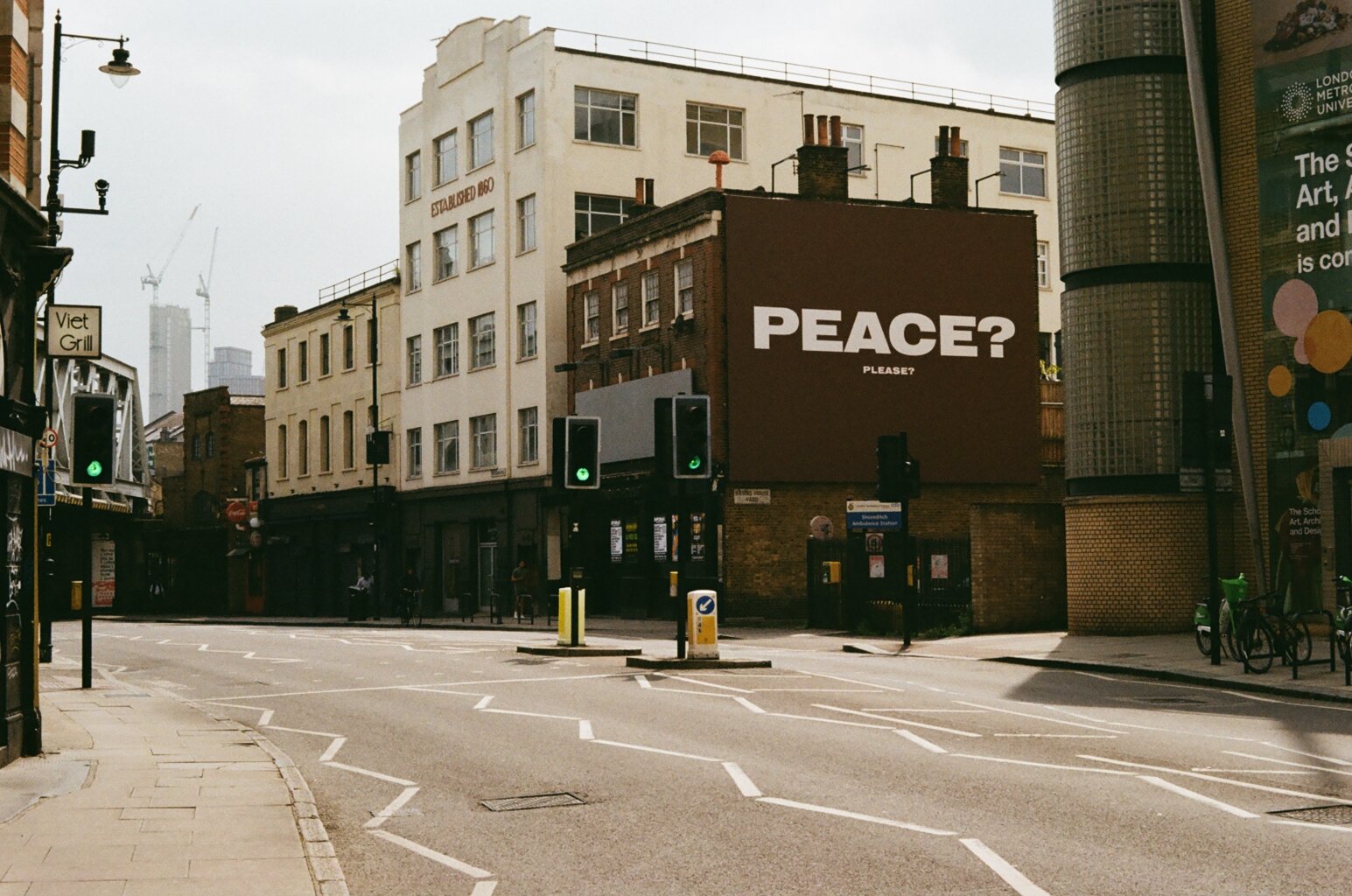 City street with traffic lights, buildings, and a large billboard that says 'PEACE? PLEASE?' in bold letters.