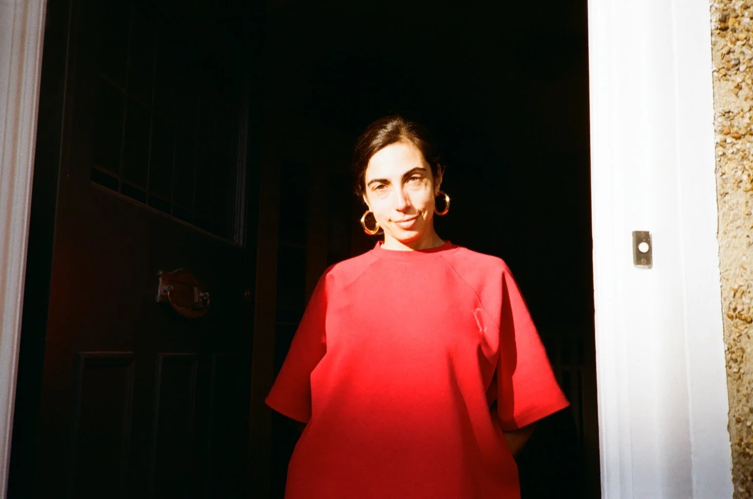 Woman with short dark hair, wearing a bright red t-shirt and gold hoop earrings, standing in a doorway with sunlight on her face.