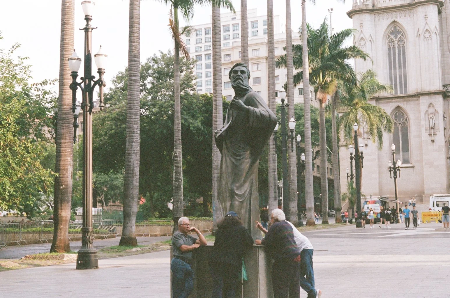 A bronze statue of a historical figure in a city park, surrounded by tall palm trees, with a group of people gathered around its base. A church is visible in the background.