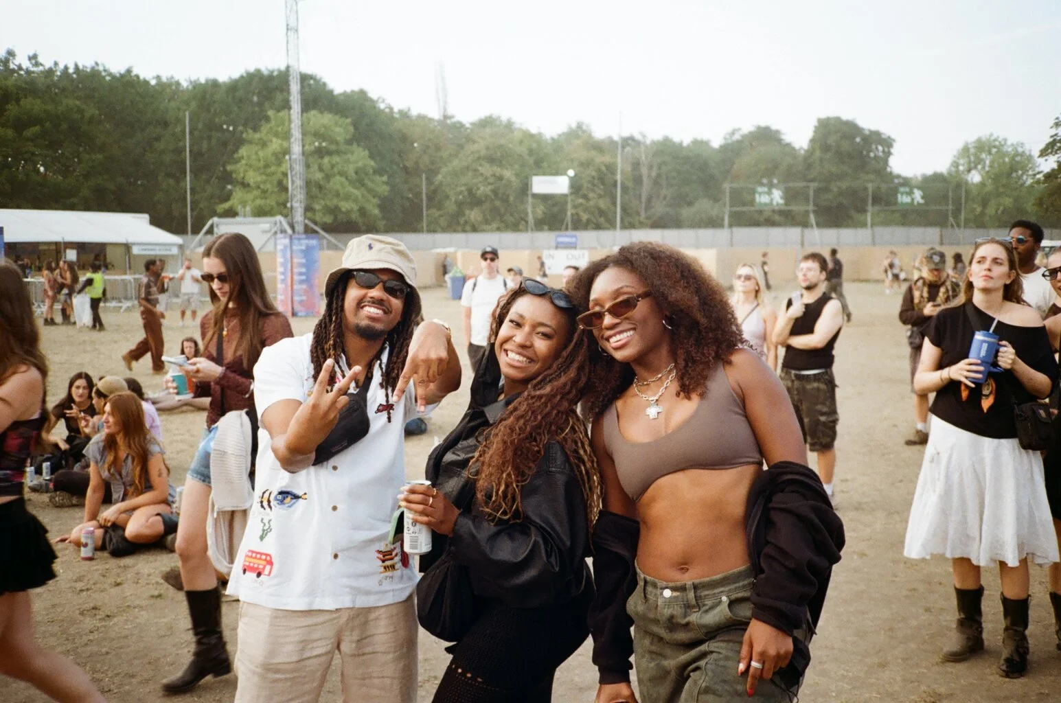 Group of three smiling friends at outdoor event, with other people in the background, some sitting on the ground and others standing, on a dirt field with trees and fences.