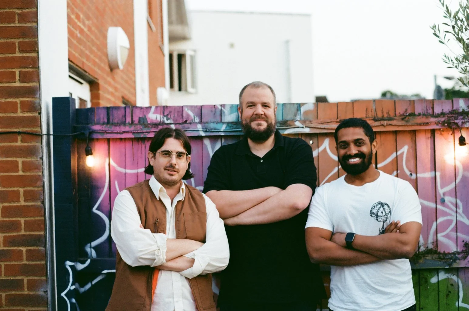 Three men standing outdoors in front of a colorful painted wooden fence, with a brick building and some windows in the background. The man on the left has glasses, dark hair, and is wearing a brown vest over a white shirt. The man in the middle has a