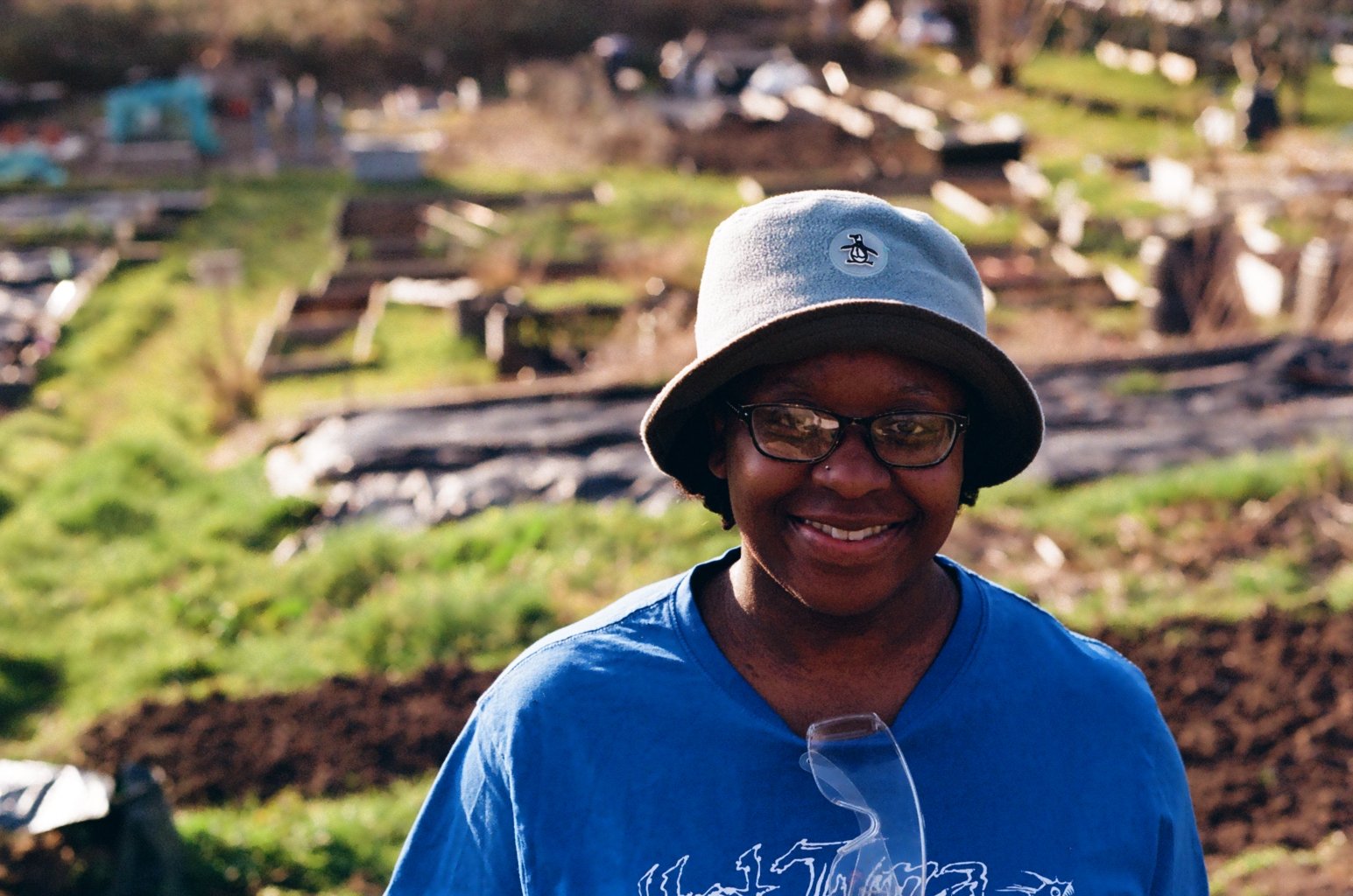 A woman with dark skin, wearing glasses, a gray bucket hat with a logo, and a blue shirt smiling outdoors during daytime. The background shows an outdoor landscape with patches of green grass and what appears to be garden beds or construction work.