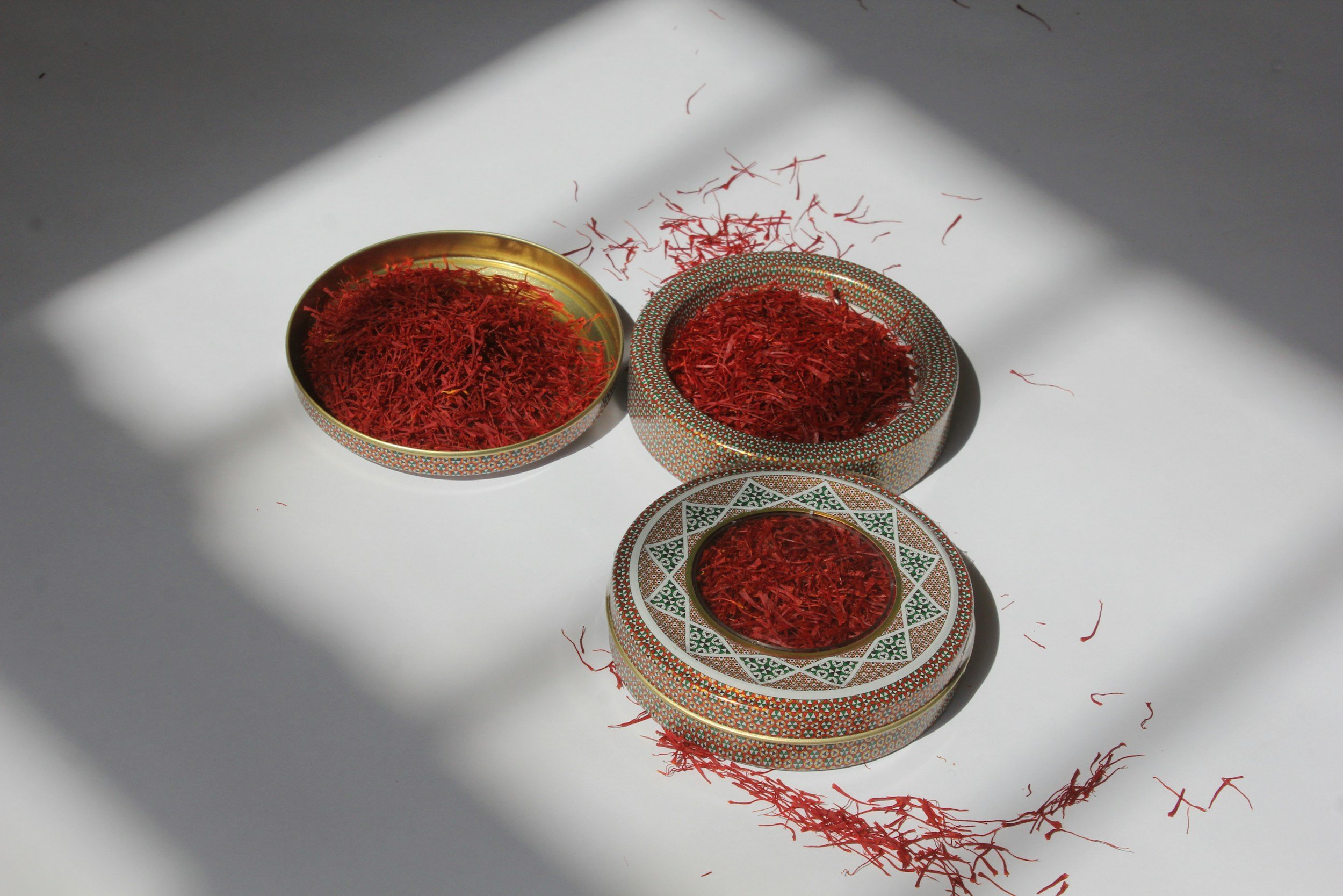 Three decorative bowls filled with red saffron threads placed on a white surface, with strands spilling out and shadows cast over the scene.