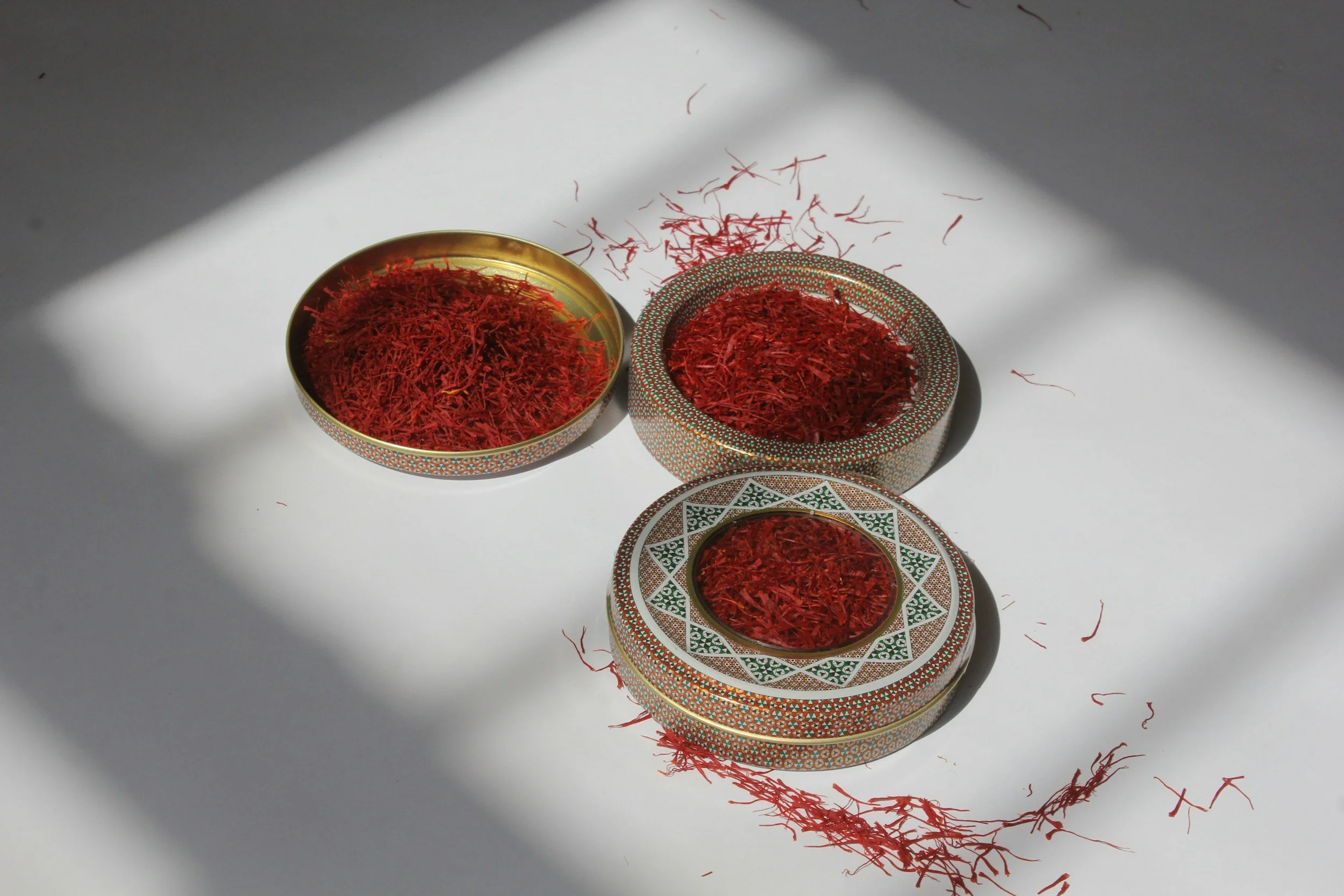 Three decorative bowls filled with red saffron threads placed on a white surface, with strands spilling out and shadows cast over the scene.