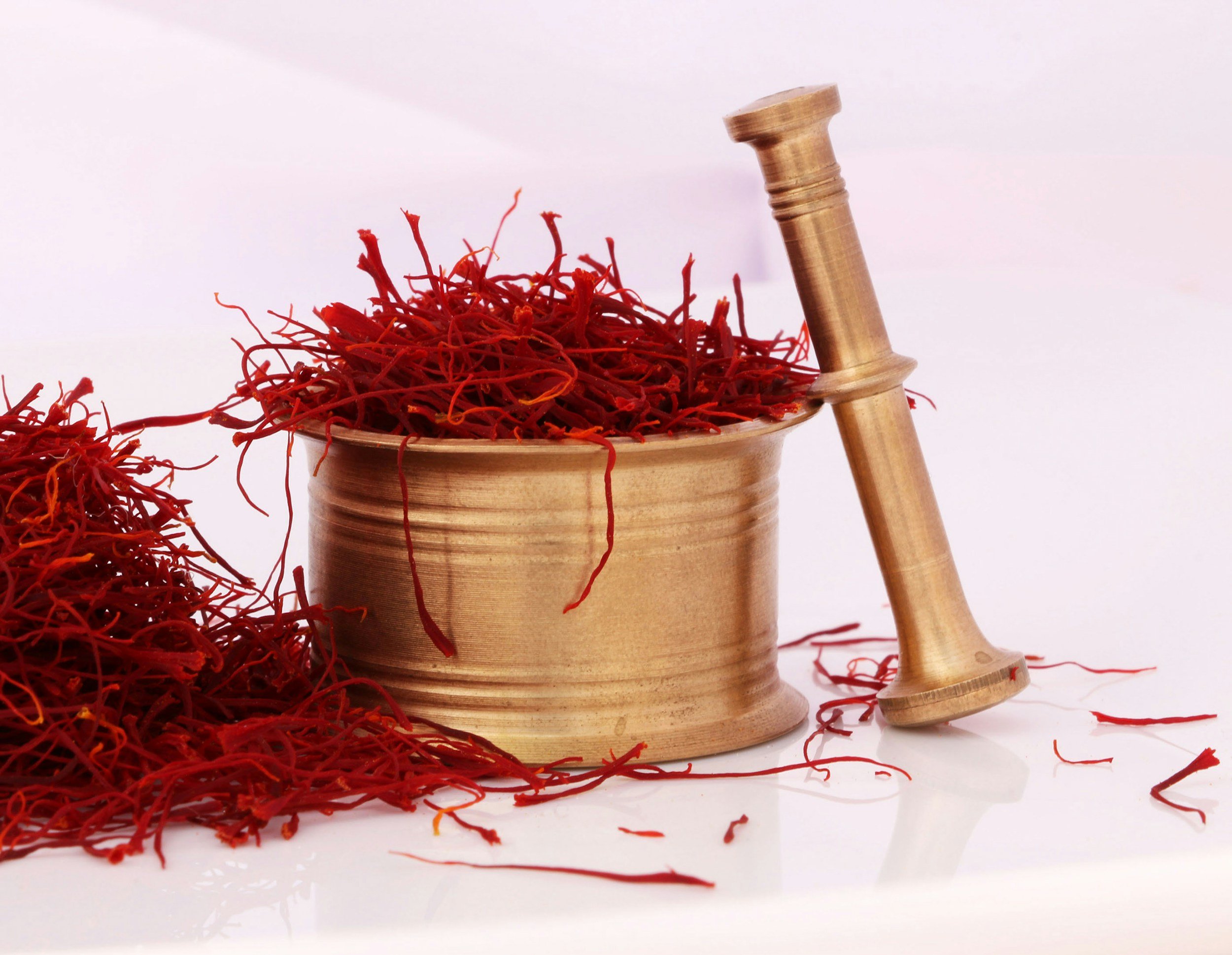 A brass mortar and pestle filled with saffron threads, with some threads spilled on a white surface.
