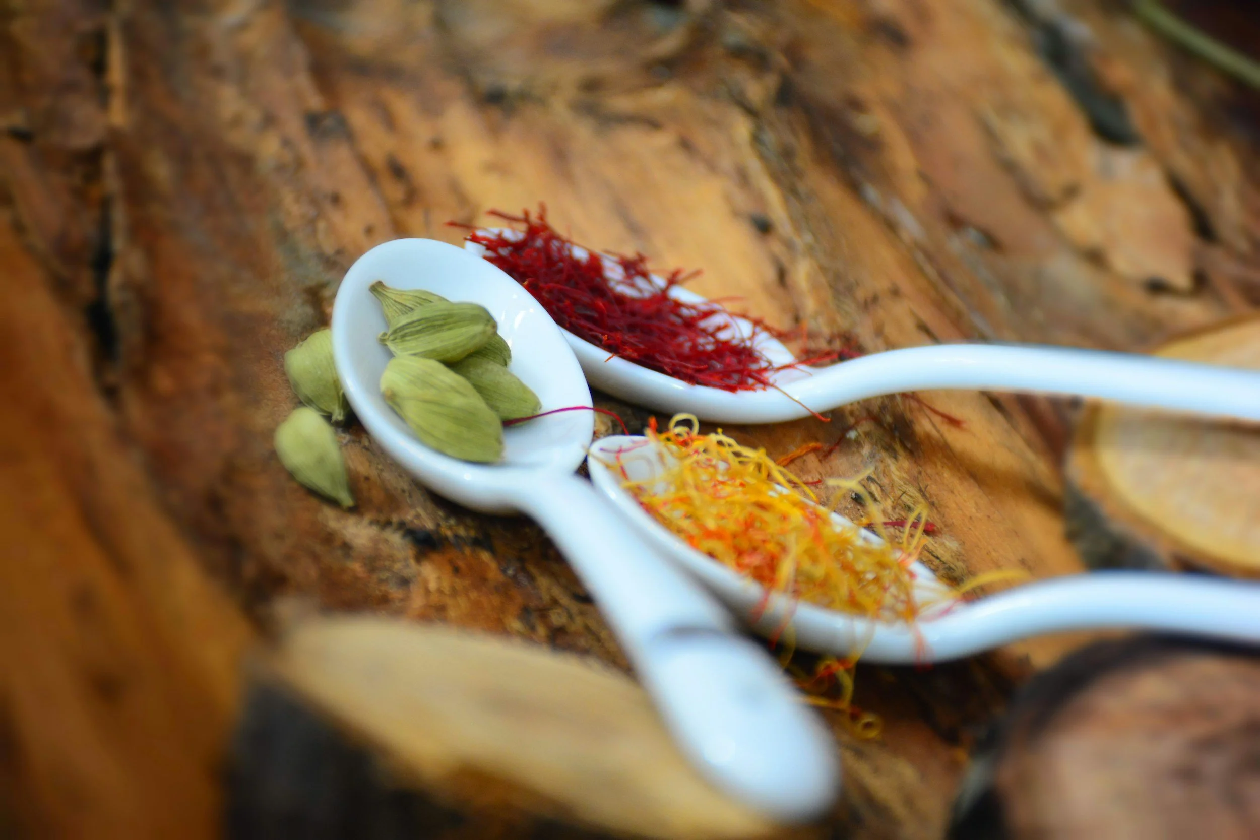 Three white ceramic spoons on a wooden surface, each filled with different colorful spices or herbs: green cardamom pods, red saffron threads, and yellow turmeric strands.