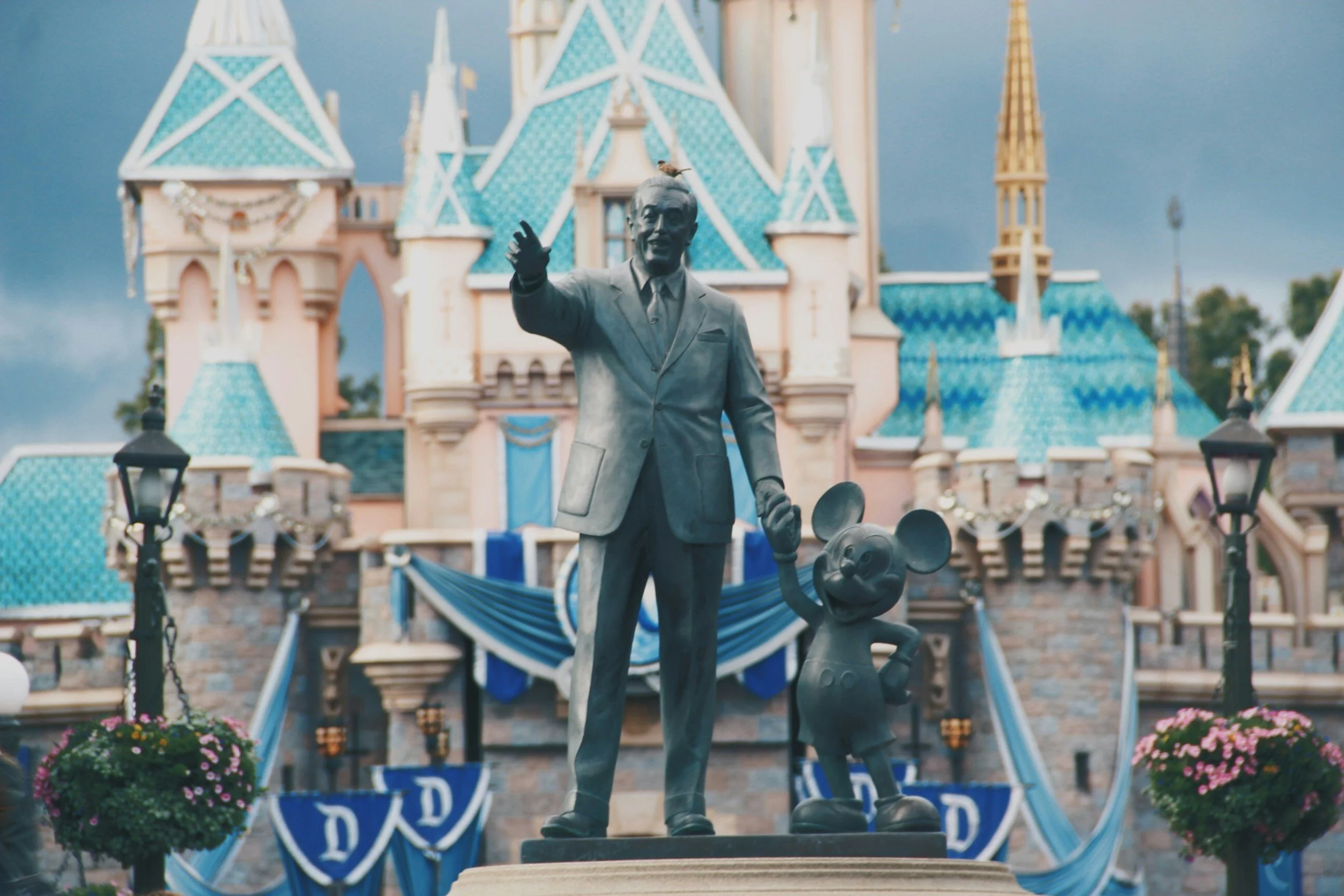 Statue of Walt Disney shaking hands with Mickey Mouse in front of Sleeping Beauty Castle at Disneyland with blue banners and flowers.