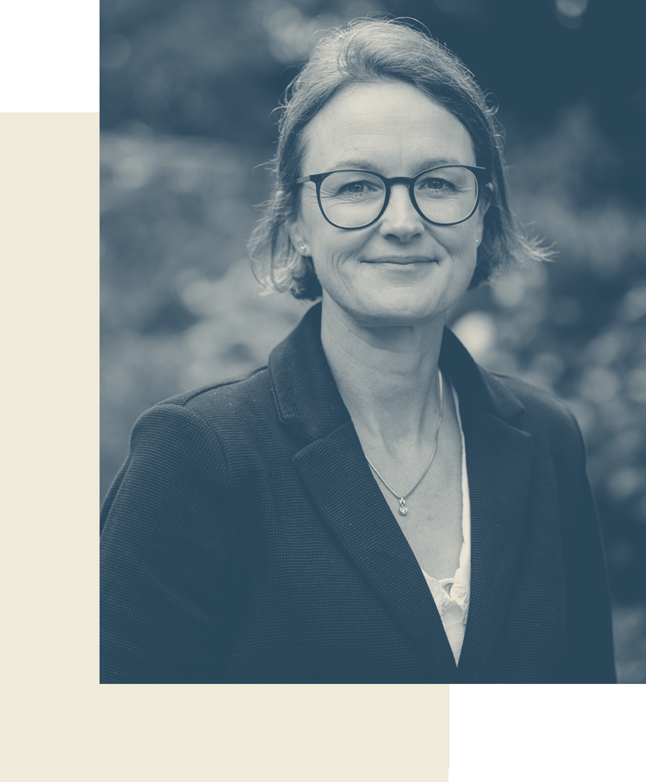 Portrait of a middle-aged woman with short hair, glasses, and a smile, wearing a dark blazer and jewelry, outdoors with blurred natural background.