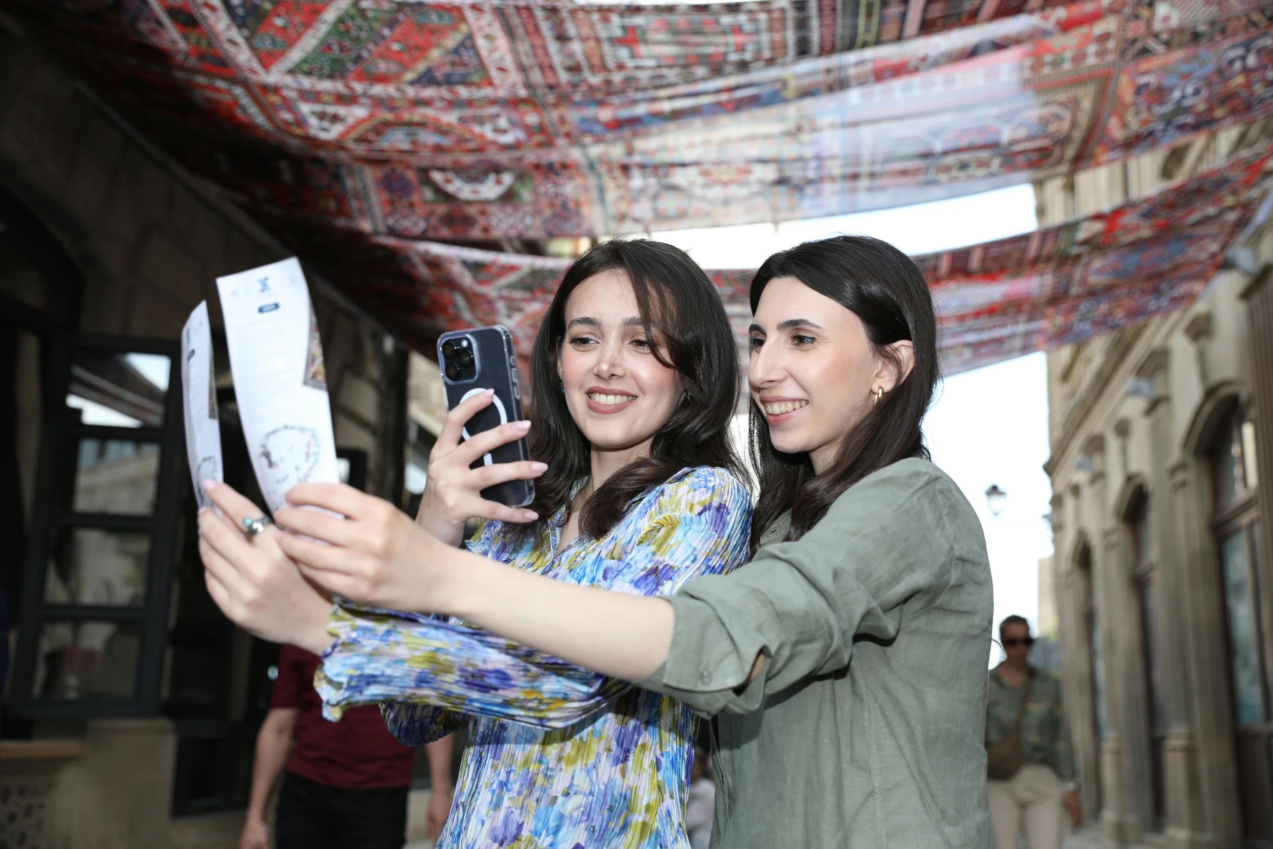 Two young women take a selfie together outdoors, with a patterned textile hanging overhead.