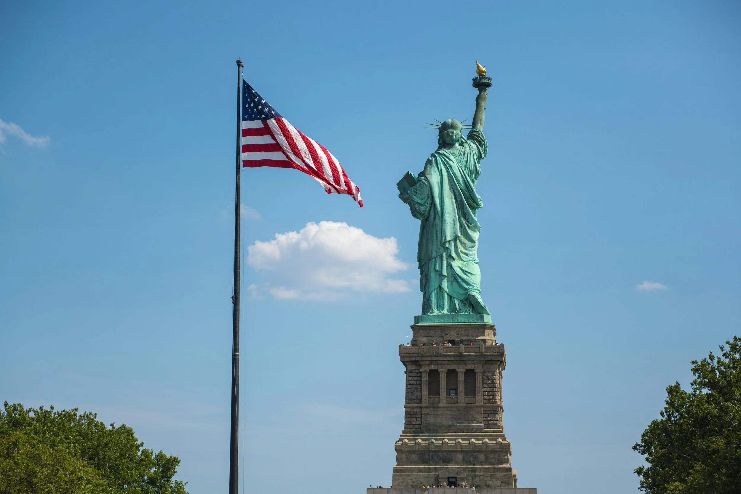 The Statue of Liberty with an American flag flying on a flagpole nearby, against a blue sky with a few clouds, and trees in the foreground.