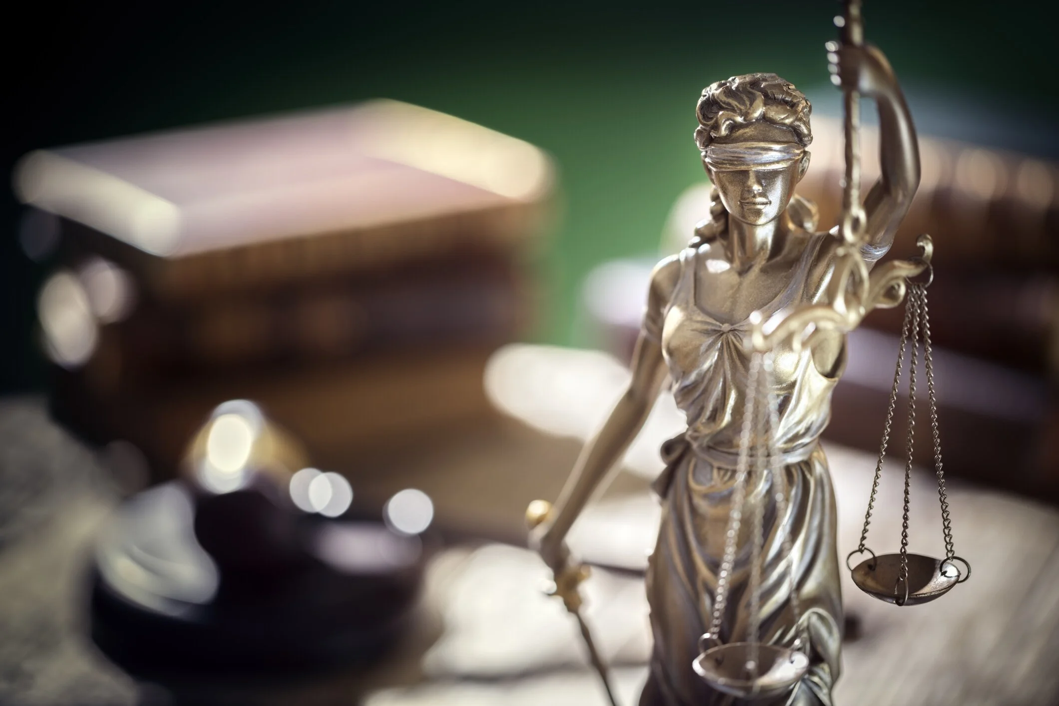 Close-up of a metallic Lady Justice statue with a blindfold, holding a sword in one hand and scales in the other, in a blurred background of legal books