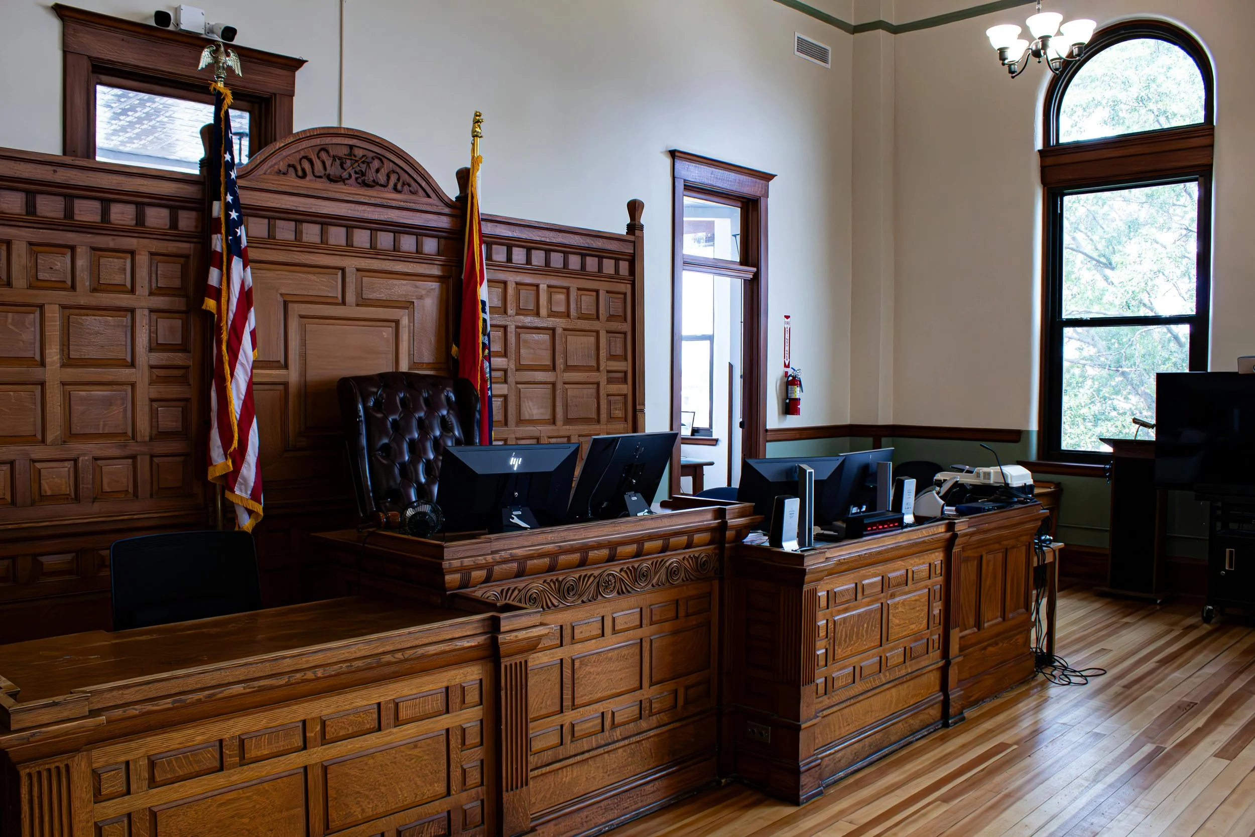 Interior of a courtroom with wooden paneling, judge's desk, computer monitors, and American flags.
