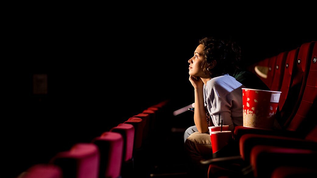 A young woman sitting alone in a movie theater, watching a film, with a large bucket of popcorn and a soda next to her.