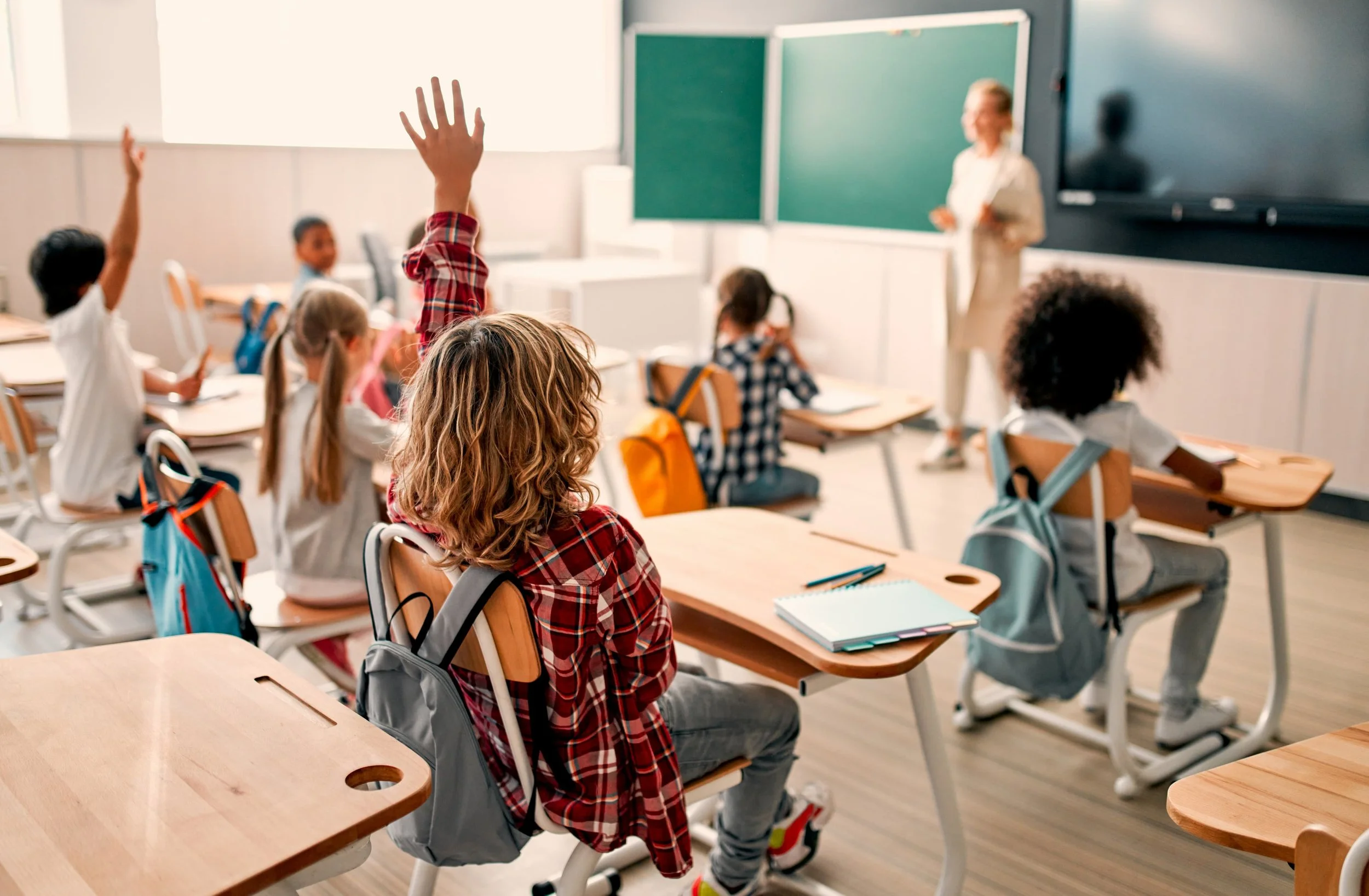 Classroom with students sitting at desks and raising hands, teacher at the front by a green chalkboard, backpacks on chairs.