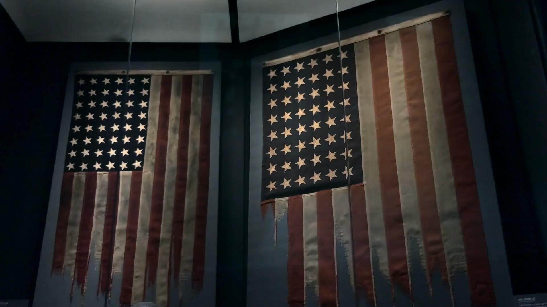 Two American flags hanging on a dark wall, with one flag appearing tattered or torn.