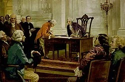 An indoor scene in a courtroom or hearing room with a judge seated at a bench, people sitting in the audience, and a woman reading or speaking from a lectern.