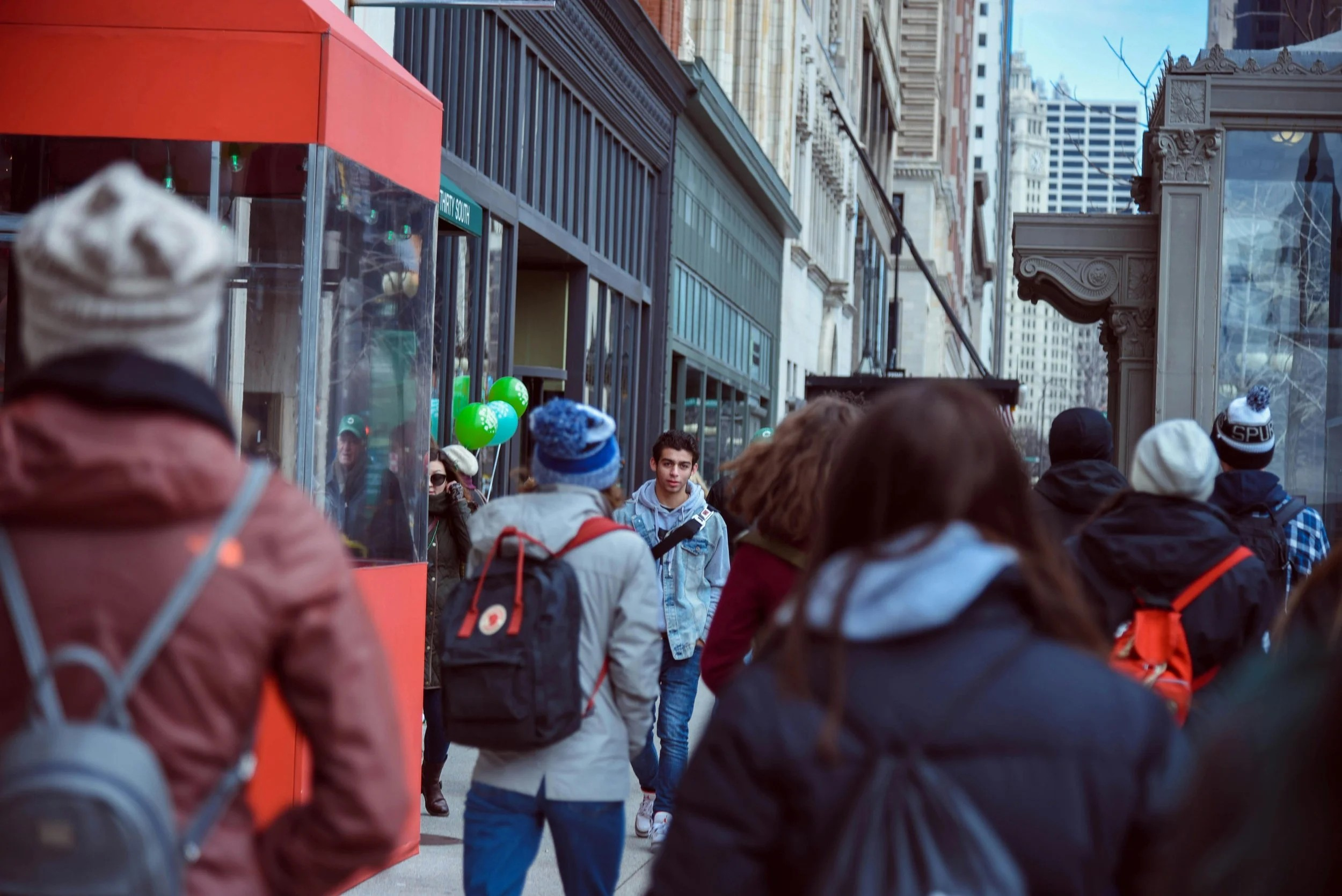 Crowd of people walking on city sidewalk with tall buildings and storefronts in the background.