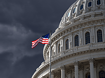United States Capitol building with an American flag flying in front under a dark, cloudy sky.